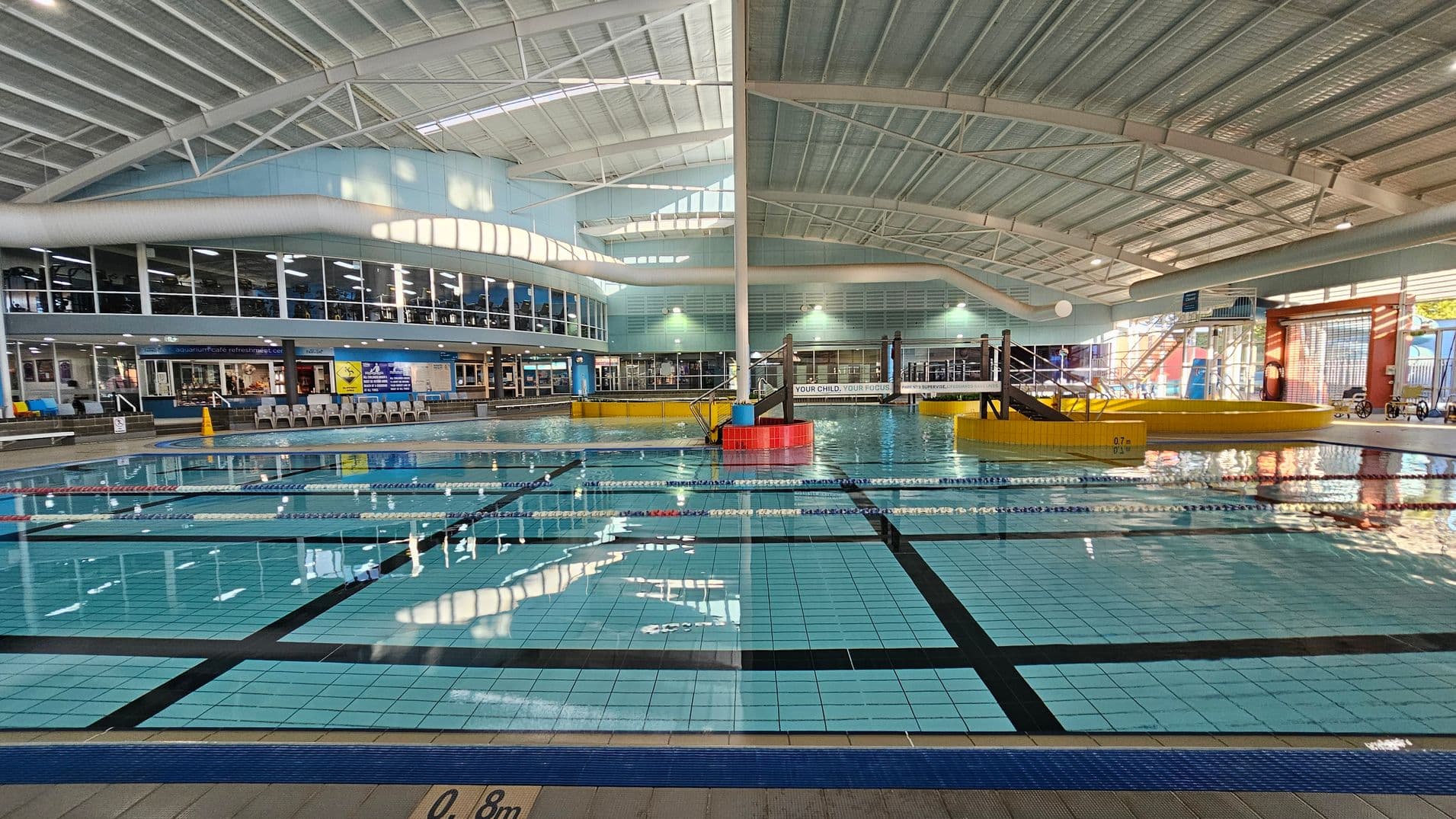 Indoor swimming pool with clear blue water and lane markings. On the left, a row of lounge chairs is visible near large windows. In the centre, a small slide and play area are surrounded by a yellow barrier. The ceiling is high with metal beams and skylights. The right side has more windows and a staircase leading to an upper level. The pool depth is marked at 0.8 metres.