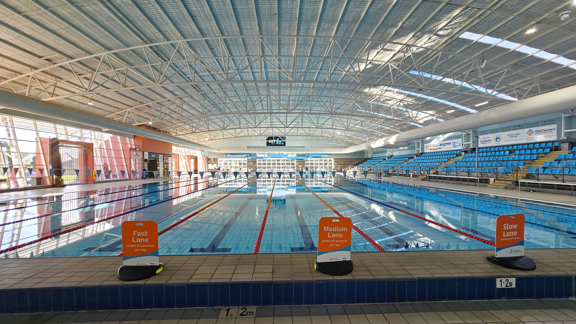An indoor swimming pool with three lanes marked by signs: "Fast Lane," "Medium Lane," and "Slow Lane." The pool is surrounded by blue seating on the right side. The ceiling is high with a metal framework and skylights. Large windows on the left allow natural light to enter. The pool deck is tiled, and the water is clear, reflecting the structure above. Signs and advertisements are visible on the walls.