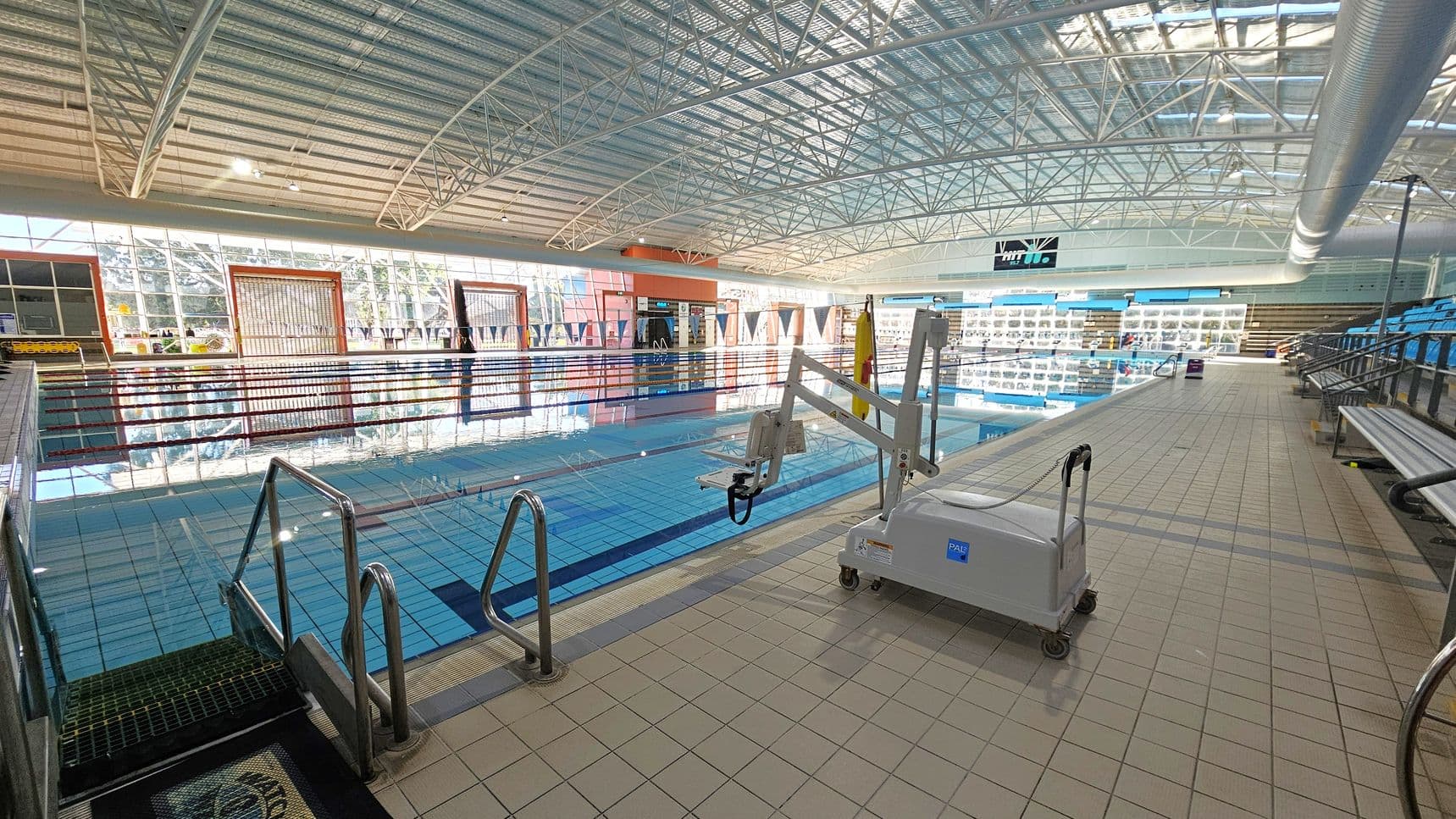 An indoor swimming pool with clear blue water and lane dividers. On the right, there's a pool lift device on the tiled deck. The left side features metal handrails leading into the pool. In the background, large windows allow natural light to illuminate the space, and there are rows of blue seating for spectators. The ceiling is high with a metal framework and a large ventilation duct running along the right side.
