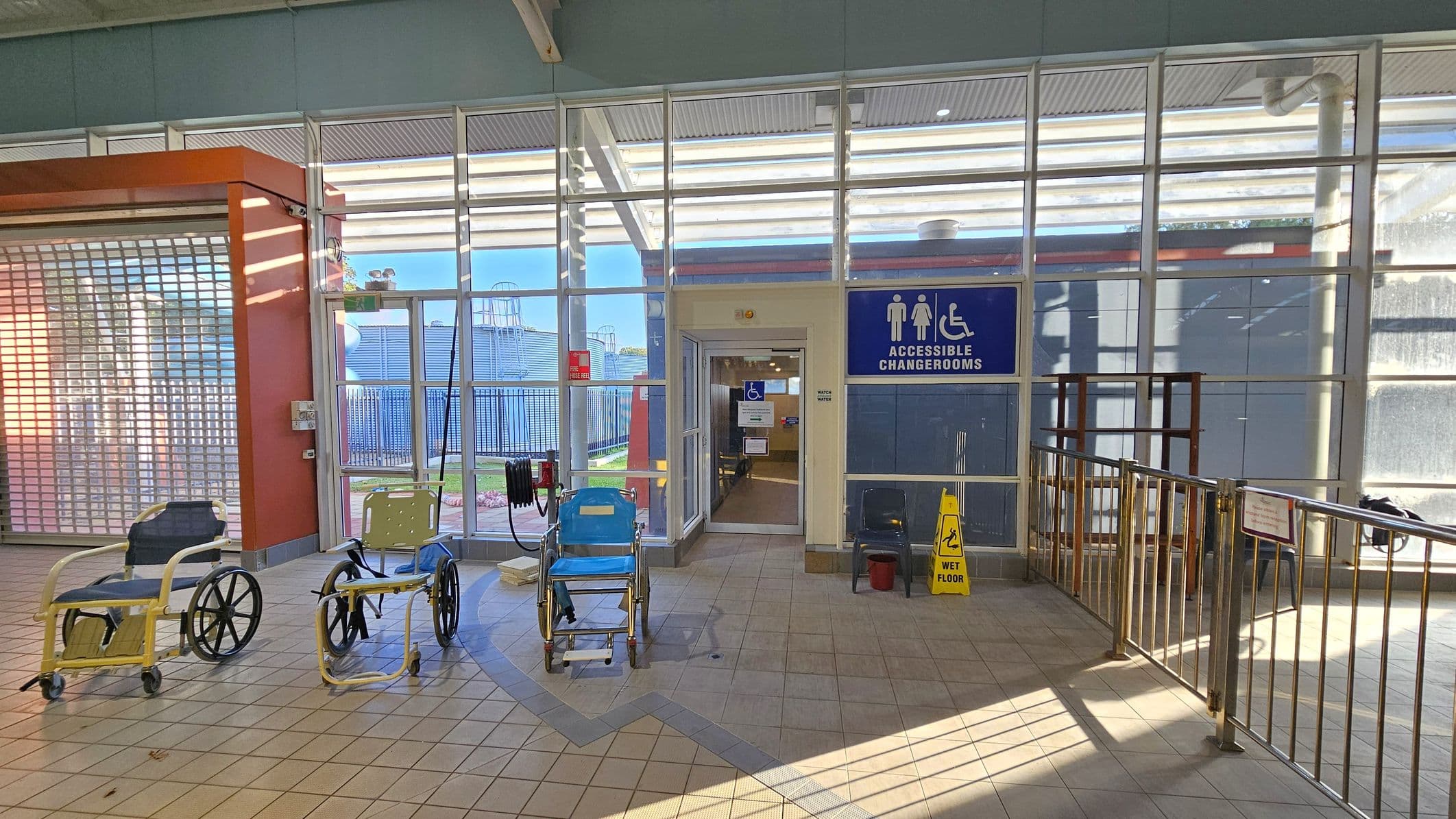 A tiled floor area with three wheelchairs positioned in front of a glass wall. The wall has a door leading to accessible changerooms, indicated by a blue sign with symbols for male, female, and wheelchair access. A yellow "wet floor" sign is placed near the entrance. To the left, there's an orange structure with a metal shutter. Metal railings are on the right side, and outside, a fenced area with some structures is visible through the glass.