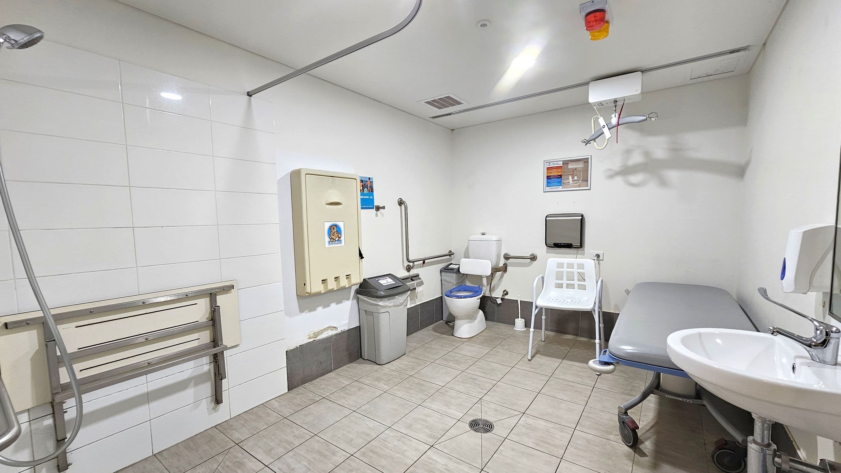 A spacious accessible bathroom with white tiled walls and a light green tiled floor. On the left, a showerhead and a foldable shower seat are mounted on the wall. In the centre, a toilet with support rails, a nappy changing station, and a waste bin are visible. On the right, a white chair, a grey padded changing table on wheels, and a sink with a soap dispenser are present. The ceiling has a hoist system and a light fixture.