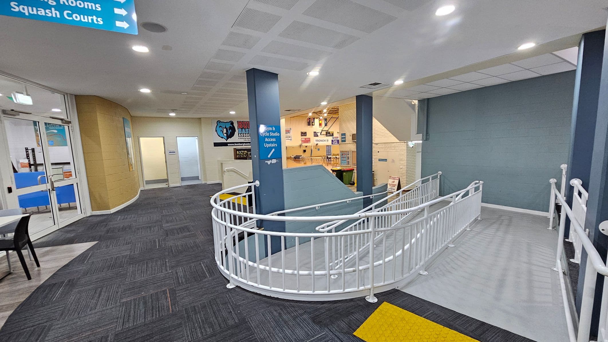 A hallway with a grey carpeted floor and a white ceiling with recessed lights. On the left, there are glass doors leading to a room with blue chairs. Signs above indicate directions to "Squash Courts" and "Change Rooms." In the centre, a blue pillar has a sign for "Gym & Cycle Studio Access Upstairs." A white railing curves around a ramp leading downwards. The walls are a mix of beige and blue, with a logo featuring a bear on the wall.