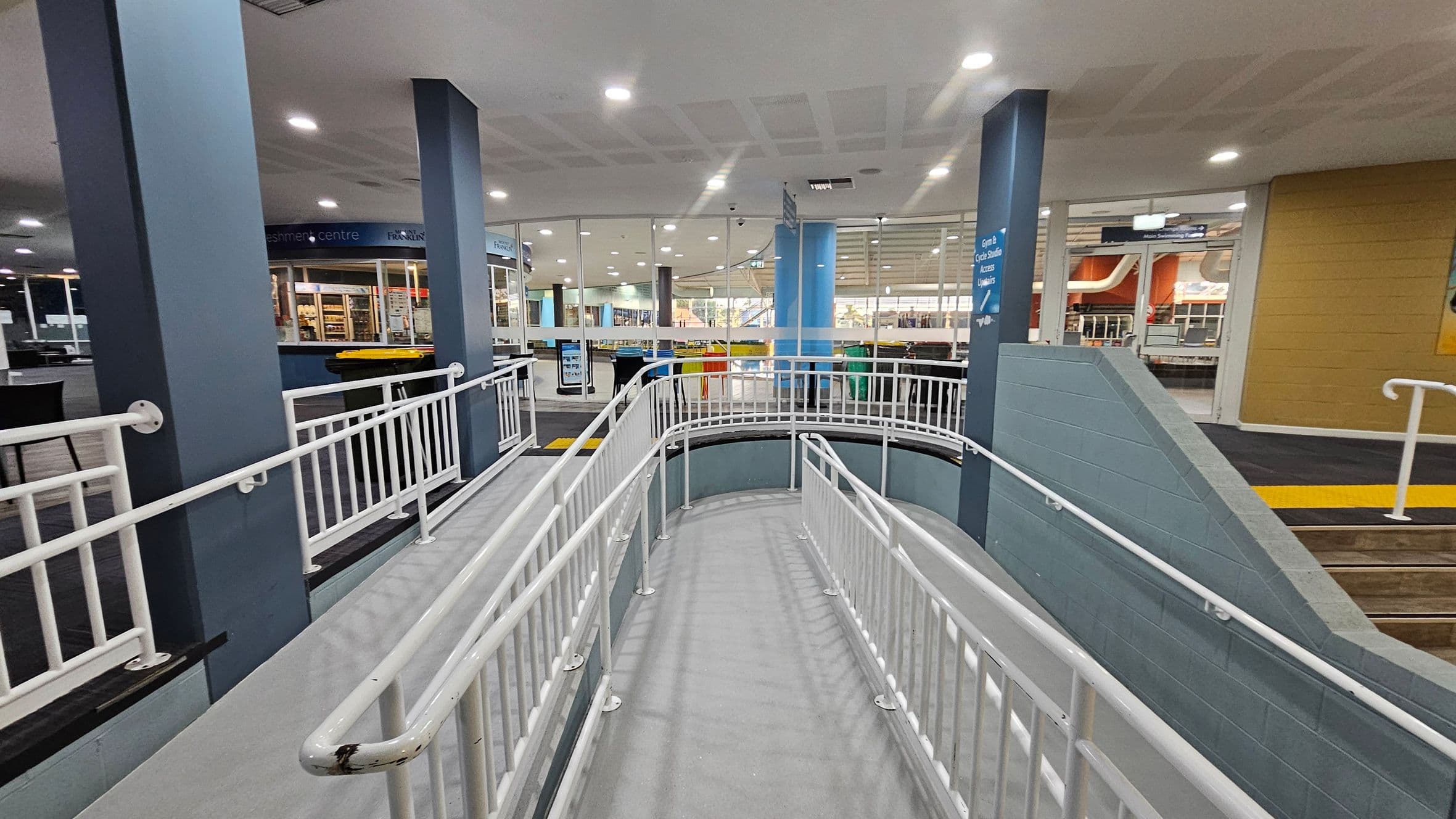 A spacious indoor area with a ramp in the centre, featuring white railings. The floor is grey, and the ceiling is white with recessed lighting. On the left, there are dark blue pillars and a refreshment centre with a counter displaying items. The right side has a yellow wall and a staircase with a yellow tactile strip. In the background, glass walls reveal more of the interior, including seating and additional structures.