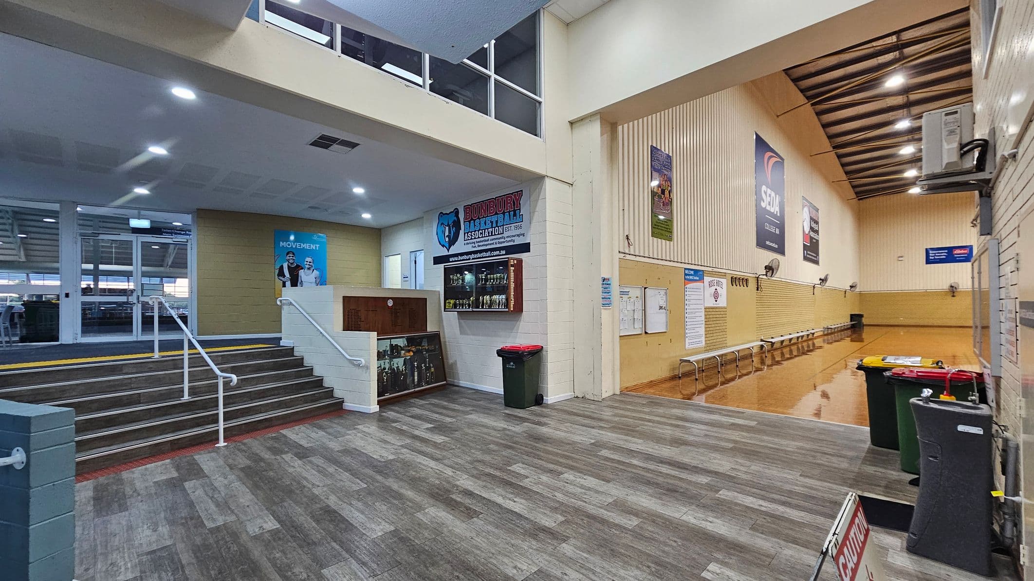 A spacious indoor area with a grey and brown tiled floor. On the left, a staircase with white railings leads up to a glass entrance. A poster with two people is visible on the wall. In the centre, a display case with trophies and a sign for a basketball association. On the right, a wooden-panelled wall with benches and several posters. The ceiling is high with exposed beams and lights. Several bins and a caution sign are in the foreground.