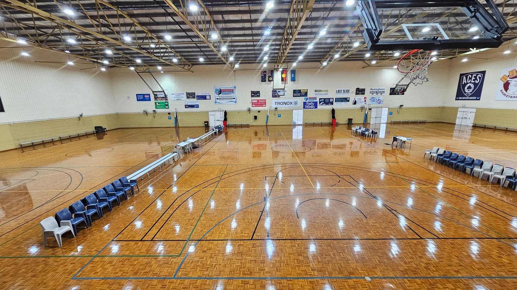 A large indoor basketball court with a polished wooden floor. On the left, there are rows of blue and white chairs along the sideline. In the centre, the court markings are visible, and a scoreboard is mounted on the far wall. On the right, more chairs are arranged, and a basketball hoop is suspended from the ceiling. The ceiling is high with exposed beams and bright overhead lights. Various banners and advertisements are displayed on the walls.