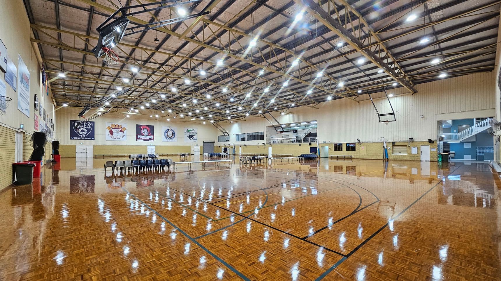A spacious indoor sports hall with a polished wooden floor marked for basketball. On the left, there are basketball hoops and banners on the walls. In the centre, rows of black chairs are arranged, and a volleyball net is set up. The right side features more chairs and a staircase leading to an upper level. The ceiling is high with exposed beams and numerous bright lights. The walls are light-coloured, and there are additional banners and doors visible.