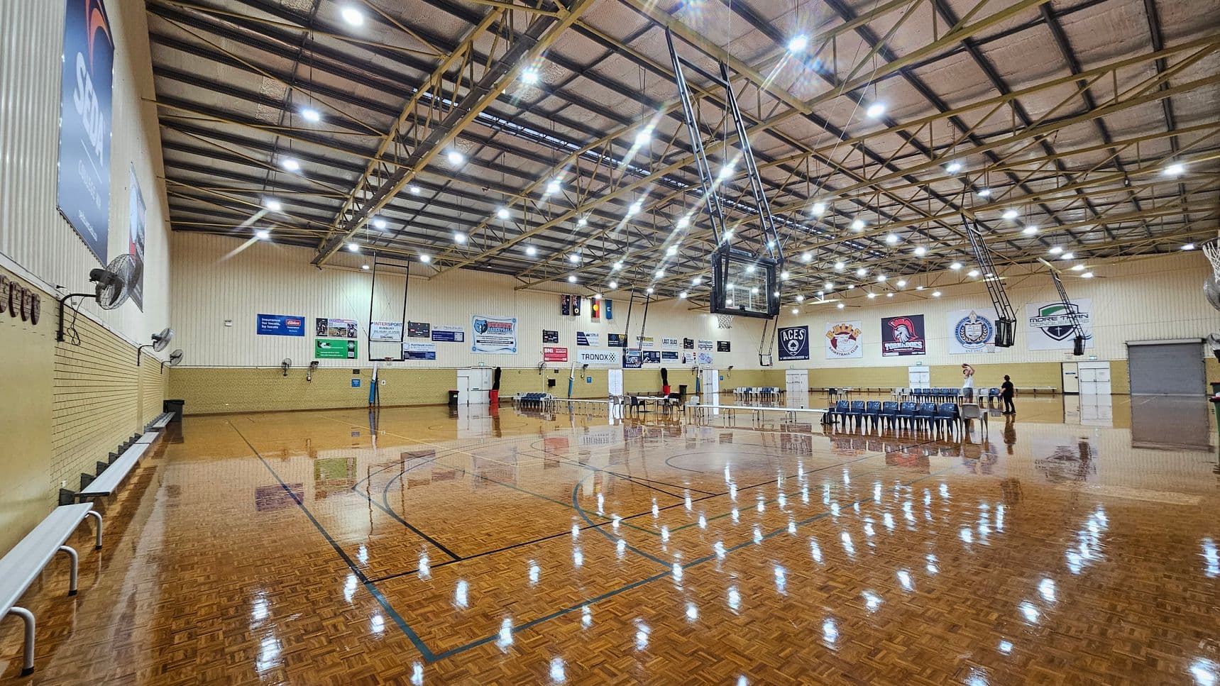 A large indoor sports hall with a polished wooden floor reflecting the overhead lights. On the left, there are benches against a brick wall with several large fans mounted above. The ceiling is high with exposed beams and numerous lights. In the centre, a basketball hoop is suspended. On the right, there are rows of stacked chairs and a few people standing near tables. The walls are adorned with various banners and logos.