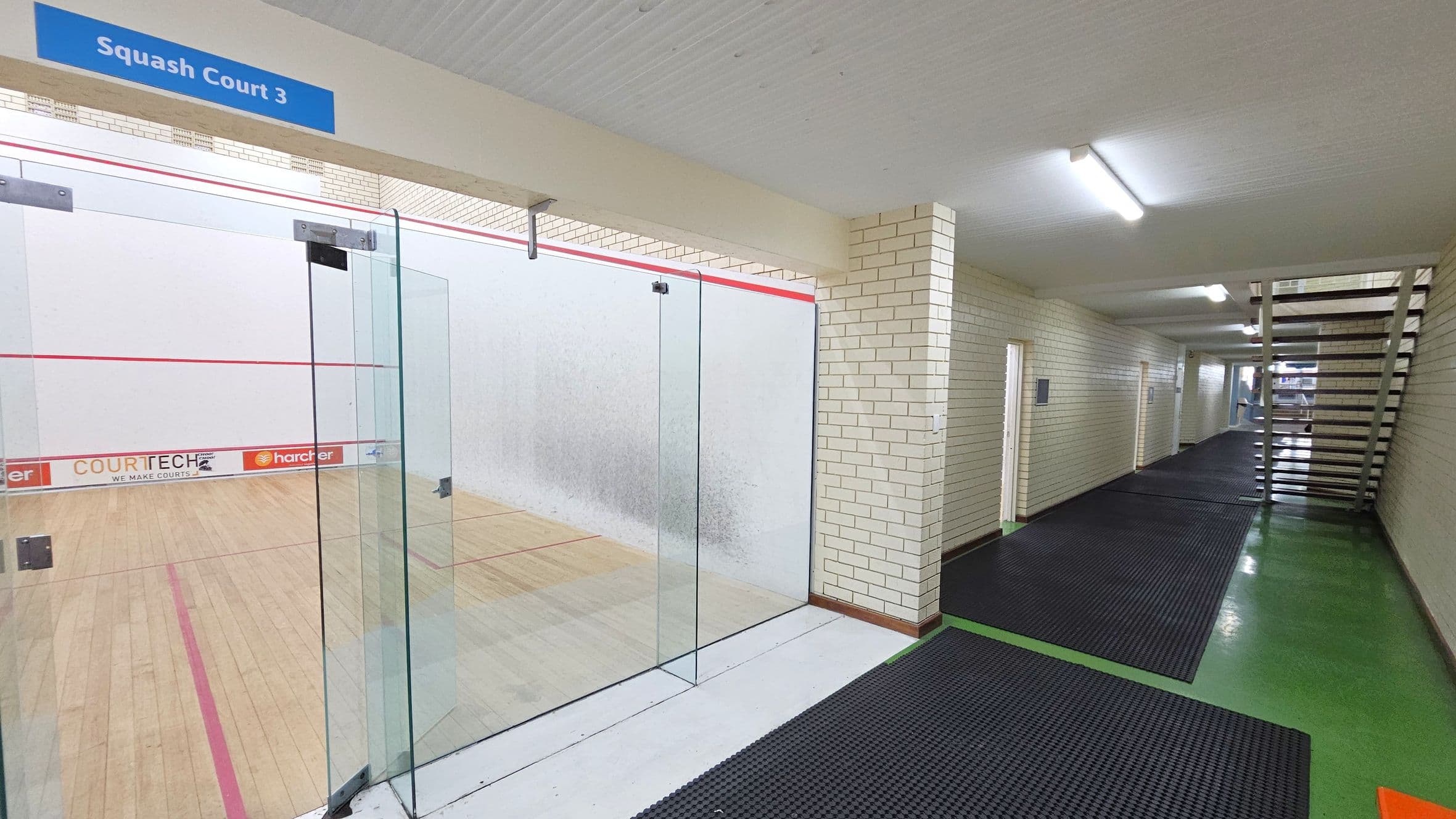 A squash court is on the left, enclosed by glass walls with a sign above reading "Squash Court 3". The court has a wooden floor with red boundary lines. On the right, a corridor with cream brick walls and black rubber mats on a green floor leads to a staircase. The ceiling is white with fluorescent lights.