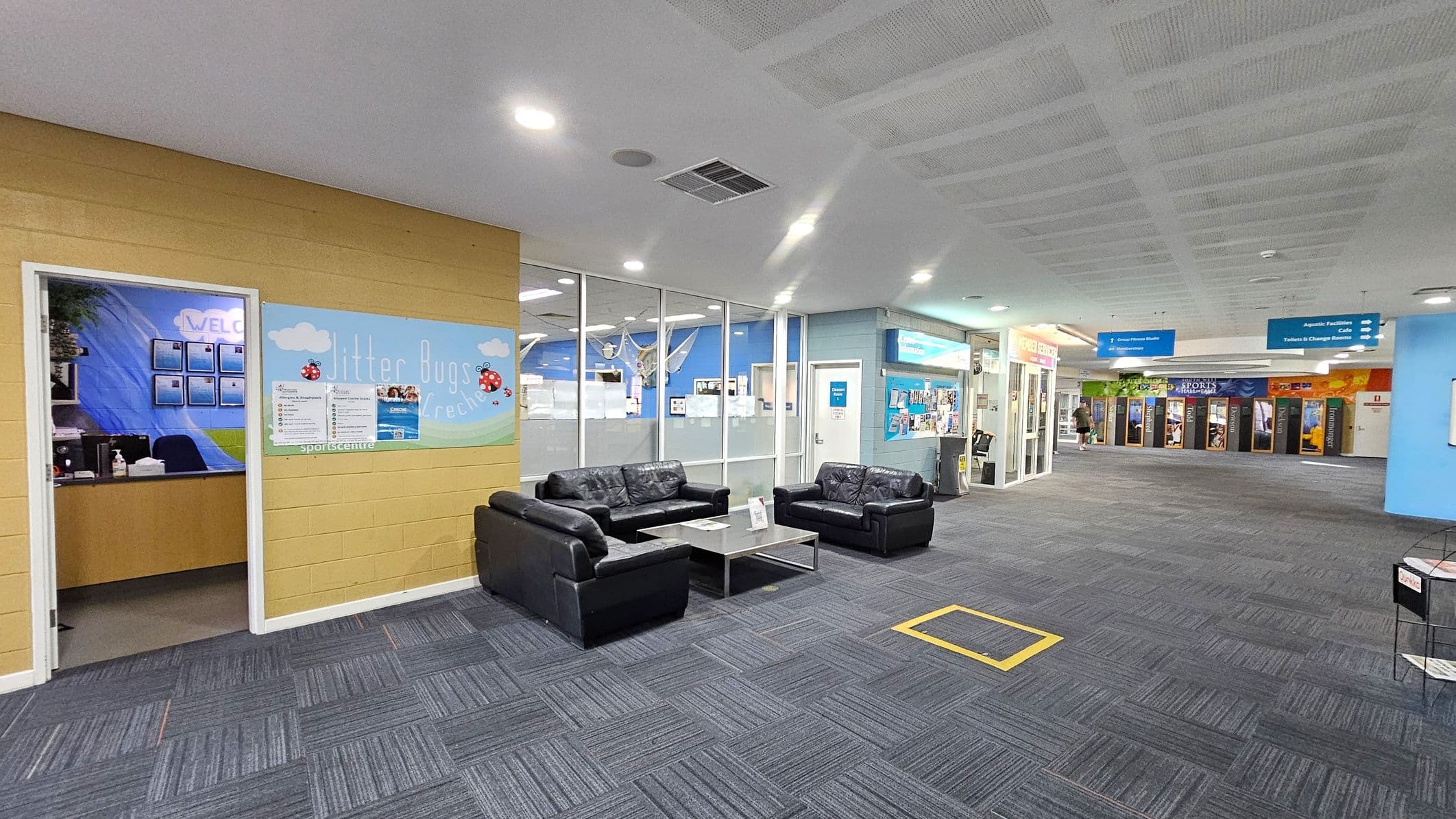 A spacious indoor area with a grey carpeted floor and a white ceiling. On the left, there's a yellow wall with a door leading to an office, featuring a sign with ladybirds. In the centre, a seating area with black leather sofas and a coffee table. The right side has glass walls and doors leading to various facilities, with signs for aquatic facilities and a café. The background shows a corridor with colourful posters and a person walking.