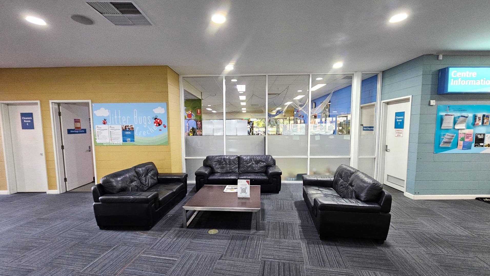A waiting area with three black leather sofas arranged around a rectangular coffee table in the centre. The floor is covered with dark grey carpet tiles. The back wall features large glass panels, providing a view into another room. On the left, there are two doors labelled "Cleaners Room 2" and "Jitterbugs Crèche" with a colourful sign. On the right, a wall-mounted display titled "Centre Information" is visible. The ceiling is white with recessed lighting.