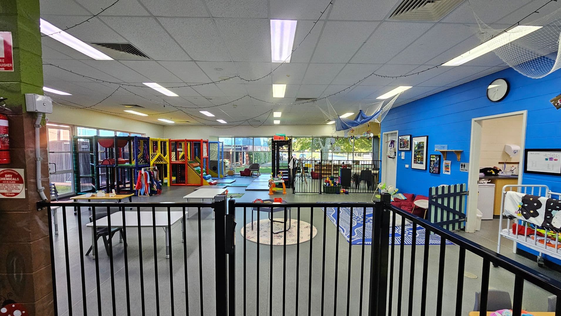 A spacious indoor play area with a grey floor and a ceiling with fluorescent lights. On the left, there are colourful play structures and a table with chairs. In the centre, a circular rug and a small play area are visible. On the right, a blue wall features framed pictures and a clock, with an open door leading to a small room. A black safety gate runs across the foreground, and string lights are draped across the ceiling.