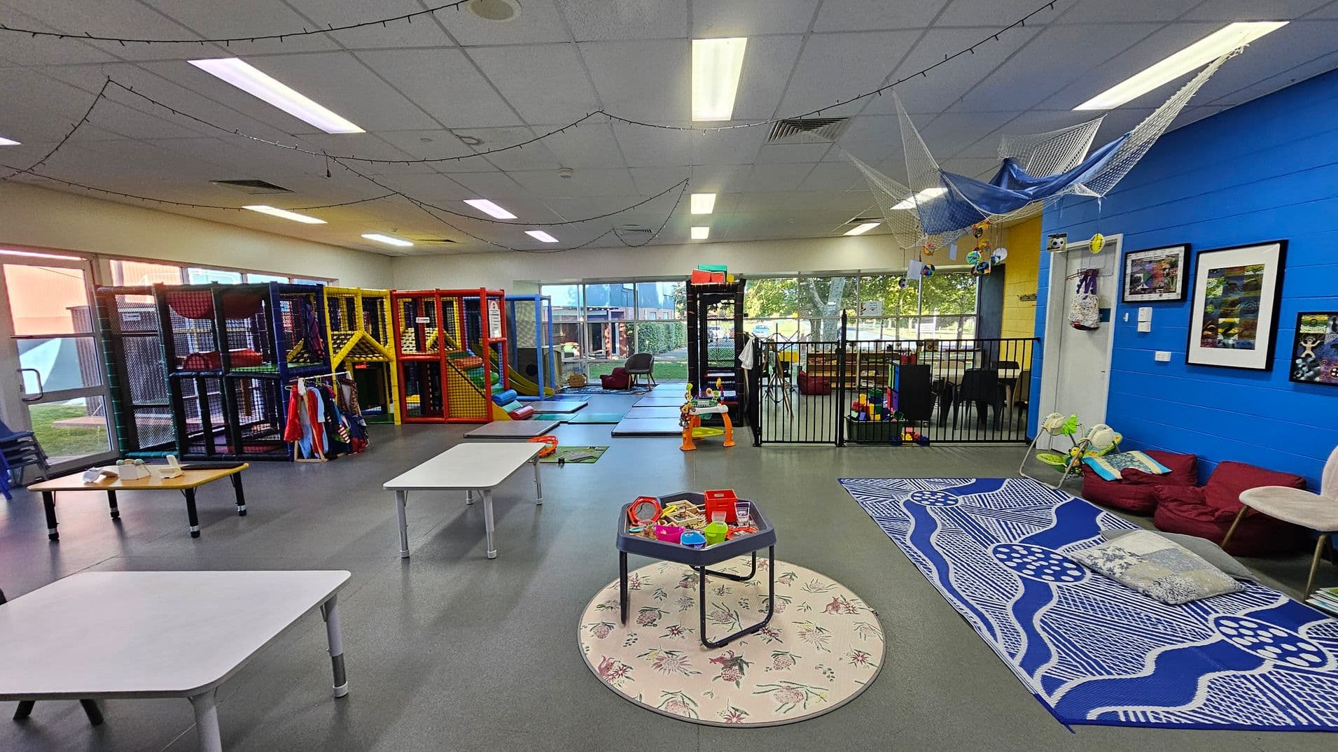 A spacious indoor play area with a grey floor and white ceiling. On the left, there are colourful play structures with slides and climbing areas. In the centre, a small round table with toys sits on a patterned rug. On the right, a blue wall features framed pictures, and a seating area with cushions and a patterned mat. The background has large windows with a view of trees outside, and a fenced-off section with more toys and seating. String lights hang from the ceiling.