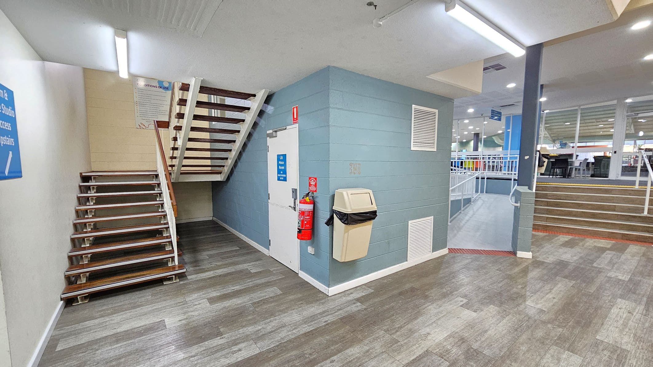A hallway with a staircase on the left leading upwards, featuring wooden steps and white railings. In the centre, a blue wall with a white door, a fire extinguisher, and a beige bin attached. The floor is covered with grey and green patterned tiles. On the right, another set of stairs with a metal railing leads to an open area with tables and chairs. The ceiling has fluorescent lights.