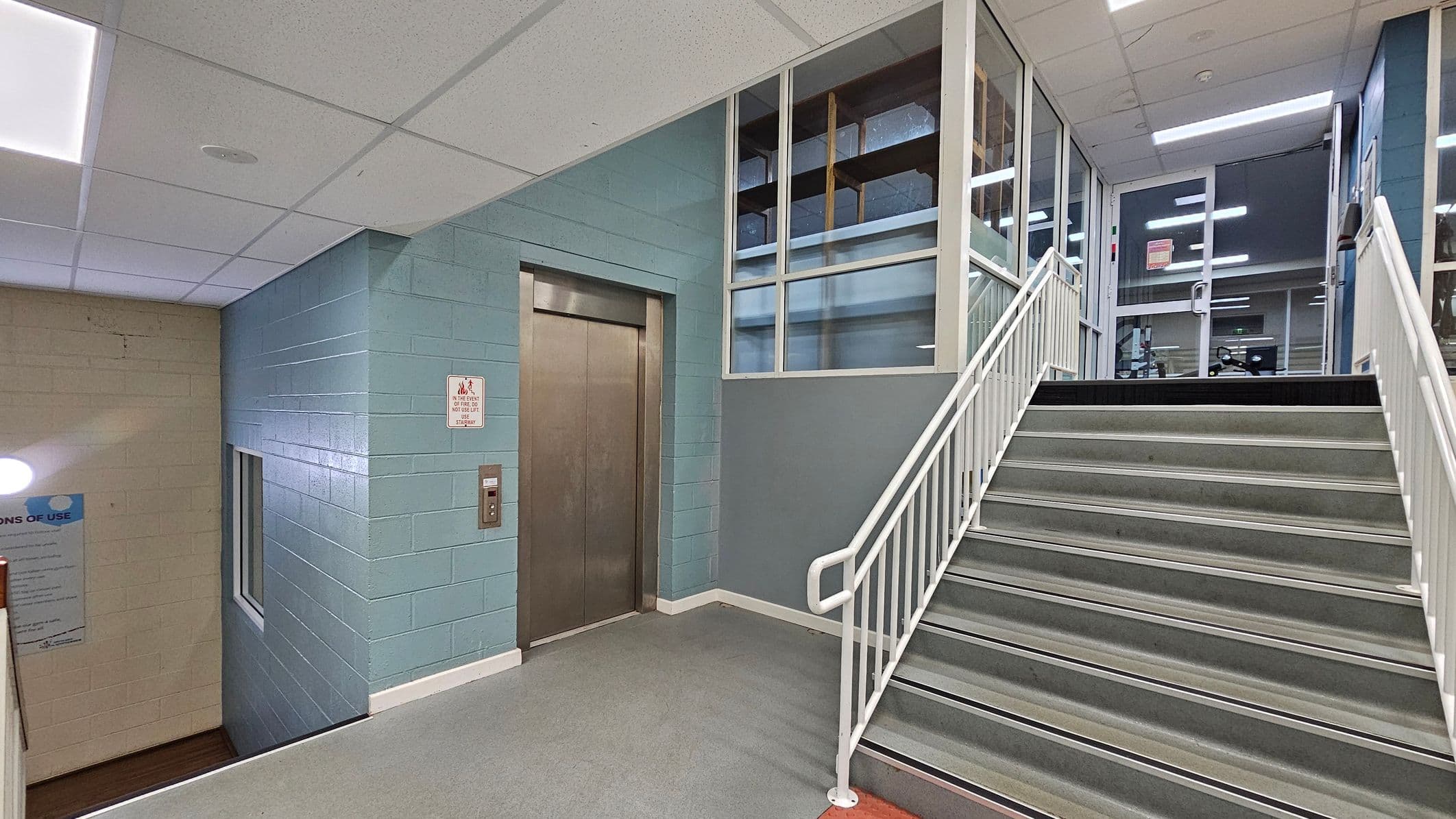A stairwell with grey steps and white railings leads upwards to a glass door. To the left, there is a metal lift door set in a light blue wall. Above the lift, there are windows with a view into another room. The ceiling is made of white tiles with fluorescent lighting. The floor is grey, and there is a sign next to the lift. A small window is visible on the left wall, which is beige.