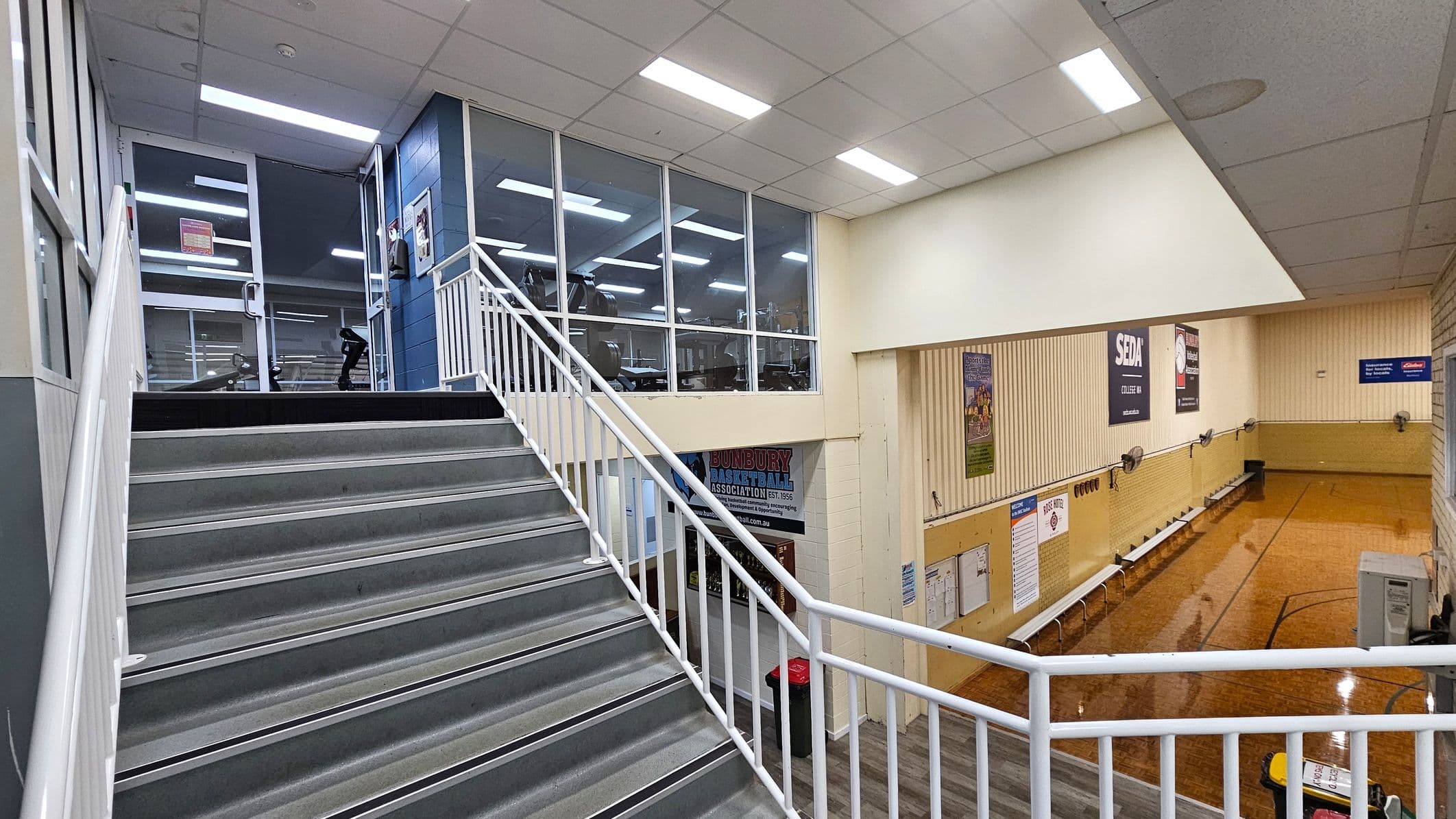 A staircase with grey steps and white railings leads up to a glass-walled area on the left, where gym equipment is visible. On the right, a basketball court with wooden flooring is partially flooded. The walls are adorned with various posters and signs. The ceiling is fitted with fluorescent lights, and the floor below the staircase is wooden.
