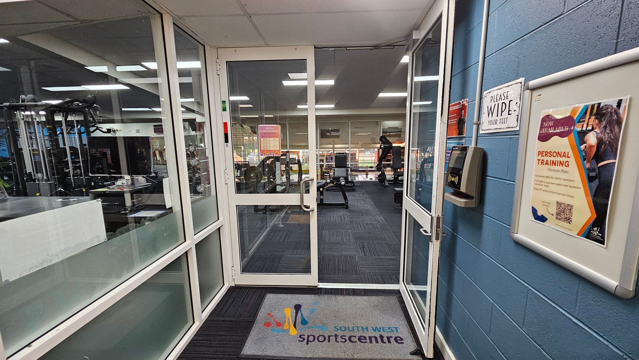 A gym entrance with glass doors and a mat on the floor labelled "South West Sports Centre". On the left, gym equipment is visible through large windows. On the right, a blue wall features a sign saying "PLEASE WIPE YOUR FEET" and a poster advertising personal training. A card reader is mounted on the wall. Inside, the gym has various exercise machines and equipment, with a carpeted floor and a ceiling with fluorescent lights.