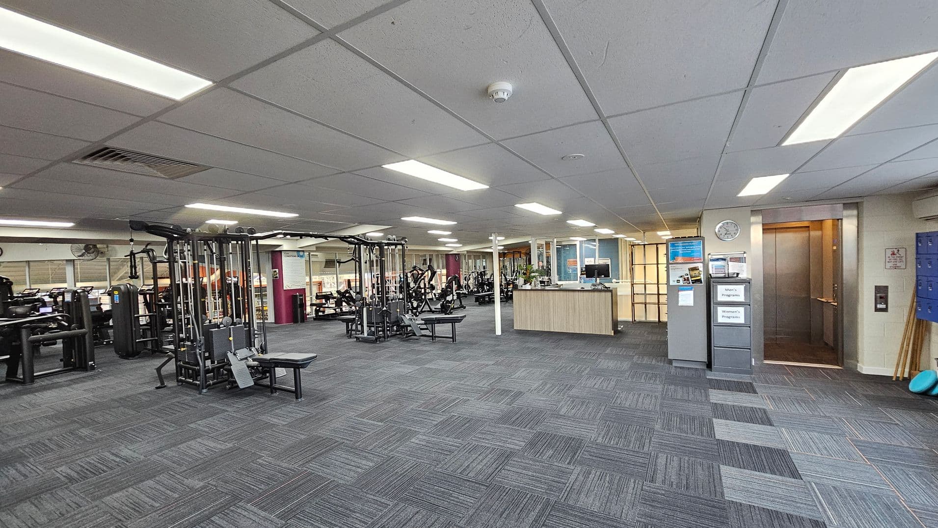 A spacious gym interior with a grey carpeted floor and a white panelled ceiling with fluorescent lights. On the left, there are various exercise machines, including weightlifting equipment. In the centre, a wooden reception desk is visible. On the right, there is an open lift with a clock above it and a set of blue lockers nearby. The walls are light-coloured, and there are informational signs and posters displayed.
