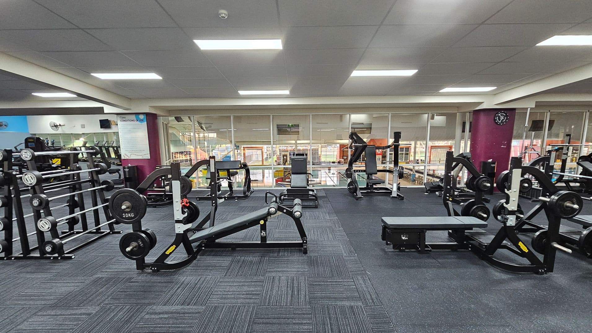 A gym with various weightlifting equipment. On the left, there are racks of dumbbells and barbells. In the centre, there are benches and weight machines. On the right, more weight machines are visible. The floor is covered with dark, patterned carpet tiles. The ceiling is fitted with fluorescent lights. The background features large windows overlooking another area of the gym, and a wall clock is mounted on a purple column.