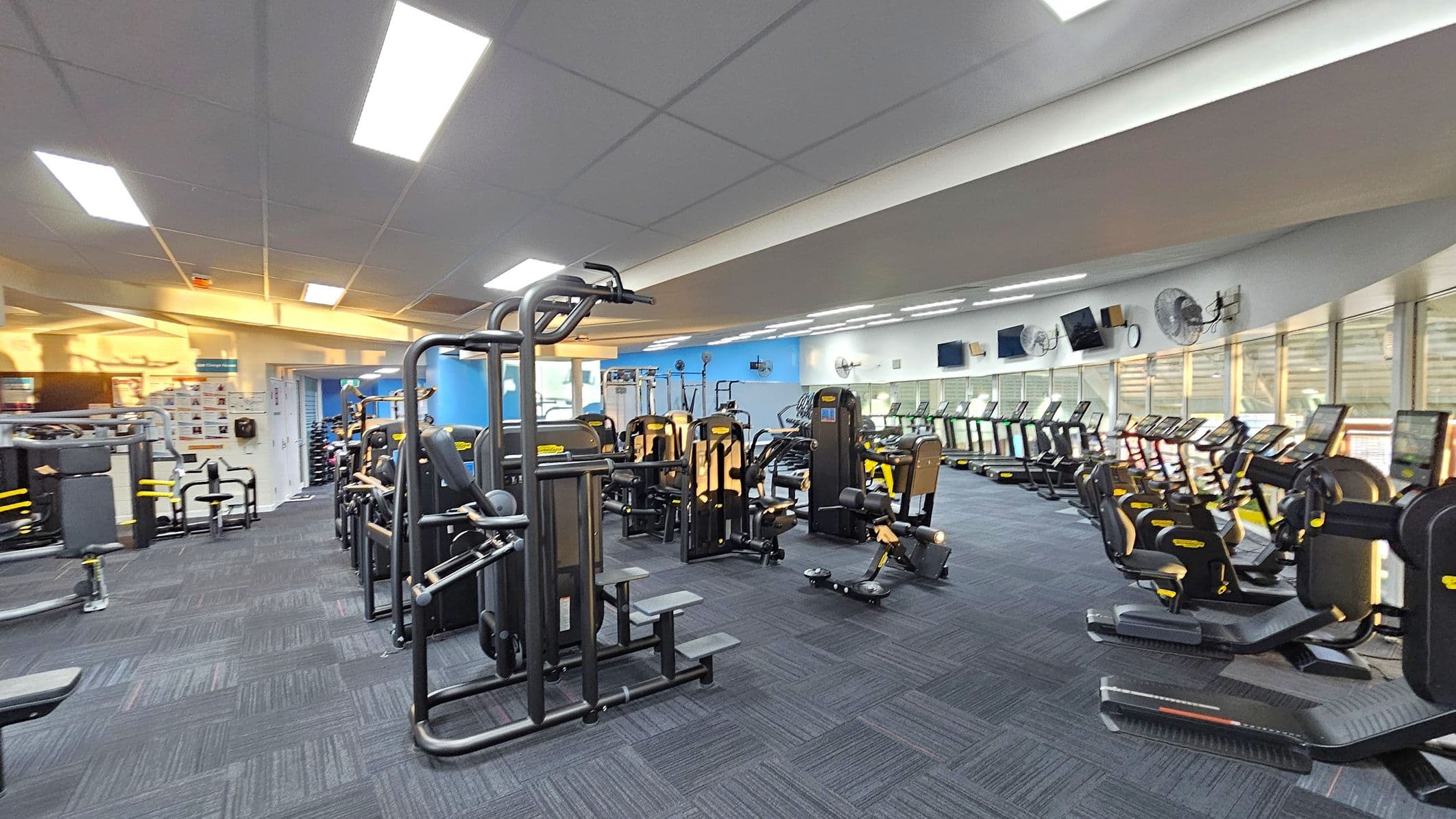 A spacious gym interior with a variety of exercise equipment. On the left, there are weight machines and benches. In the centre, more weight machines are arranged in rows. On the right, a line of treadmills and exercise bikes faces large windows. The floor is covered with dark blue carpet tiles, and the ceiling has bright fluorescent lights. The walls are painted blue and white, with several fans and televisions mounted above the treadmills.
