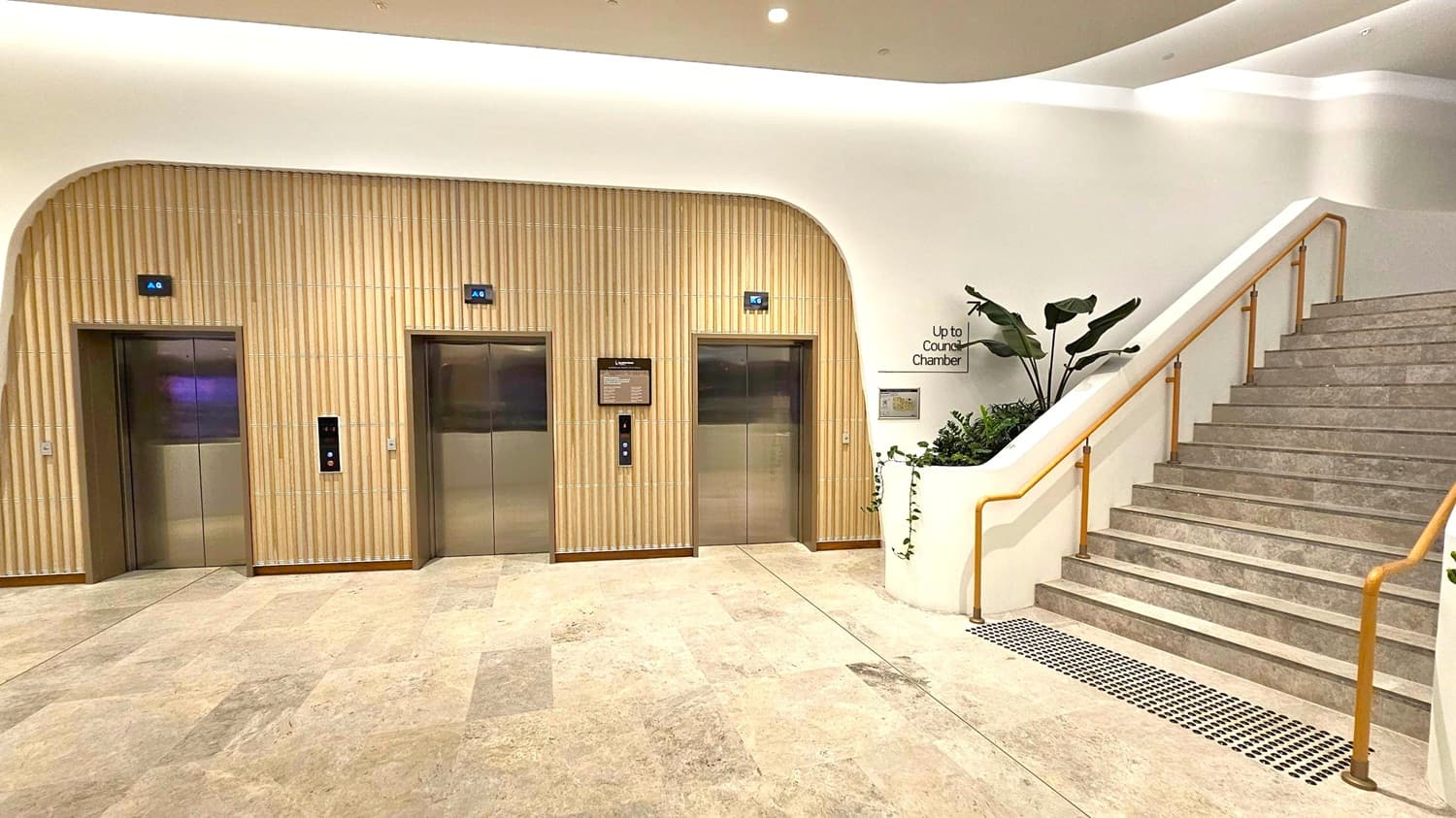 On the left, there are two lift doors with digital floor indicators above them, set in a wall with vertical wooden panelling. In the middle, a sign and lift buttons are visible between the lifts. On the right, a staircase with a wooden handrail leads upwards, accompanied by a planter with green plants. The floor is tiled in a light stone pattern, and the ceiling is smooth with recessed lighting.