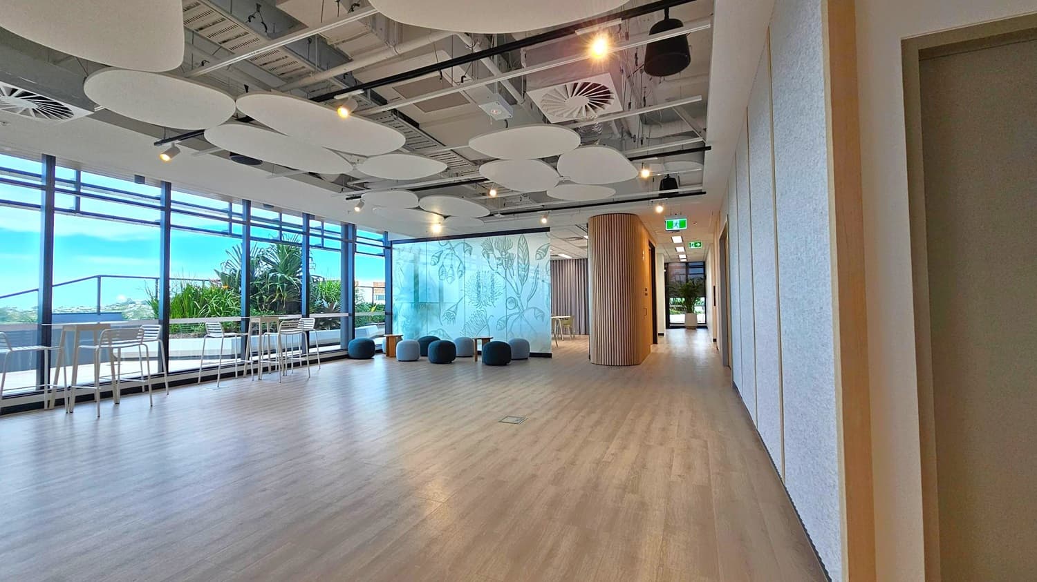 A spacious, modern interior with light wooden flooring. On the left, large windows offer a view of greenery and outdoor seating with high tables and chairs. In the centre, a decorative glass wall with botanical designs is flanked by round, cushioned stools. The ceiling features circular acoustic panels and exposed ductwork. On the right, a hallway leads to another area, with a wooden column and exit signs visible.