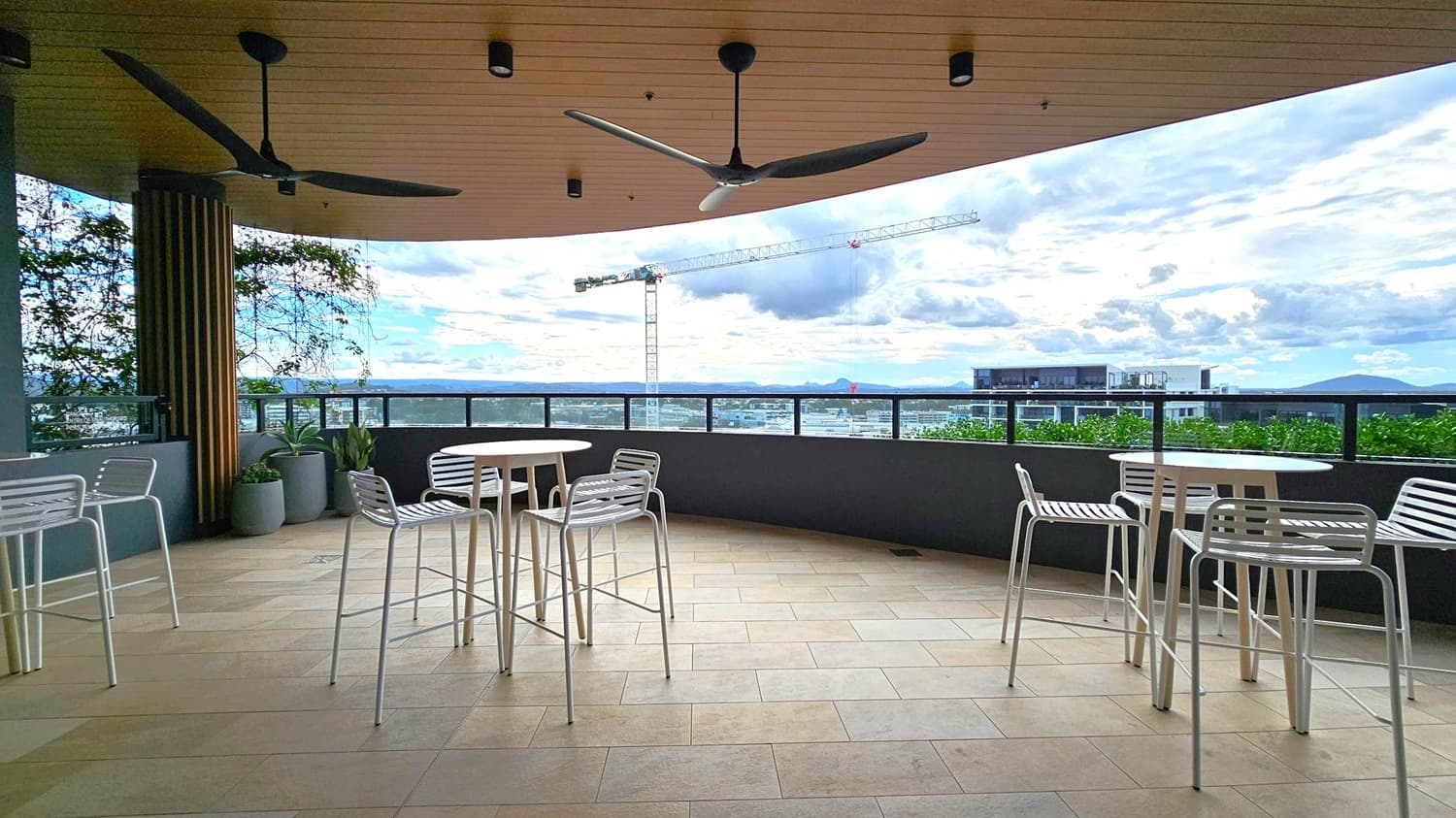 A rooftop terrace with a tiled floor and a wooden ceiling featuring two large ceiling fans. On the left, there are potted plants and a round table with white chairs. In the centre, another round table with chairs is positioned near a railing. The background shows a cityscape with a construction crane and distant mountains. The terrace is partially enclosed with a glass barrier, providing a view of the surroundings.