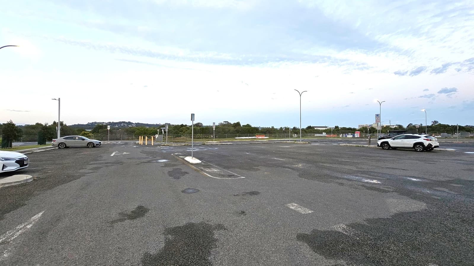 An empty car park with a few vehicles scattered around. On the left, a silver car is parked near a streetlight. On the right, a white SUV is parked under another streetlight. The ground is asphalt with visible patches and markings. In the background, there are trees and some distant buildings. Several streetlights are lit, casting a soft glow over the area. The sky is overcast with a hint of light.