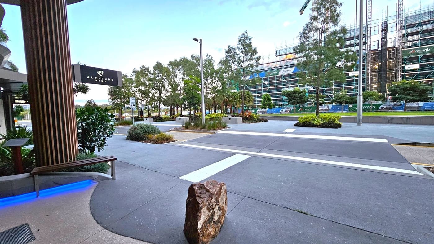 On the left, a building with a sign reading "Altitude" and a wooden column. A bench with blue lighting underneath is nearby. In the centre, a wide pavement with a large rock and a pedestrian crossing. Trees and shrubs line the area. On the right, a construction site with scaffolding and a building under development. The background features more trees and a clear sky.