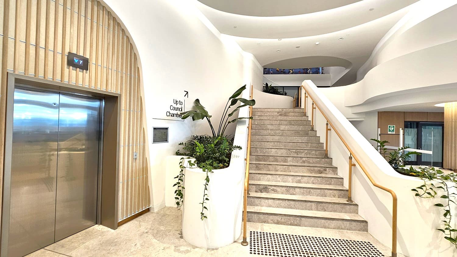 A modern interior with a staircase on the right, featuring a light stone finish and a sleek handrail. To the left, a stainless steel lift with a digital floor indicator above. Between the lift and the stairs, a planter with lush green foliage. The walls are smooth and white, with a sign indicating the direction to the council chamber. The ceiling has a curved, layered design, and the floor is a light stone with a textured mat near the stairs.