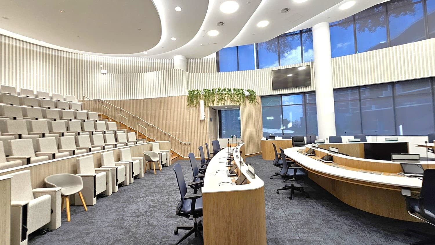 A modern conference room with tiered seating on the left, featuring light grey upholstered chairs. In the centre, there are curved wooden desks with black office chairs and computer monitors. The back wall has vertical wooden panelling and a large screen, with plants hanging above a doorway. The right side has large windows with blue-tinted glass. The ceiling is white with recessed lighting, and the floor is covered in dark grey carpet.