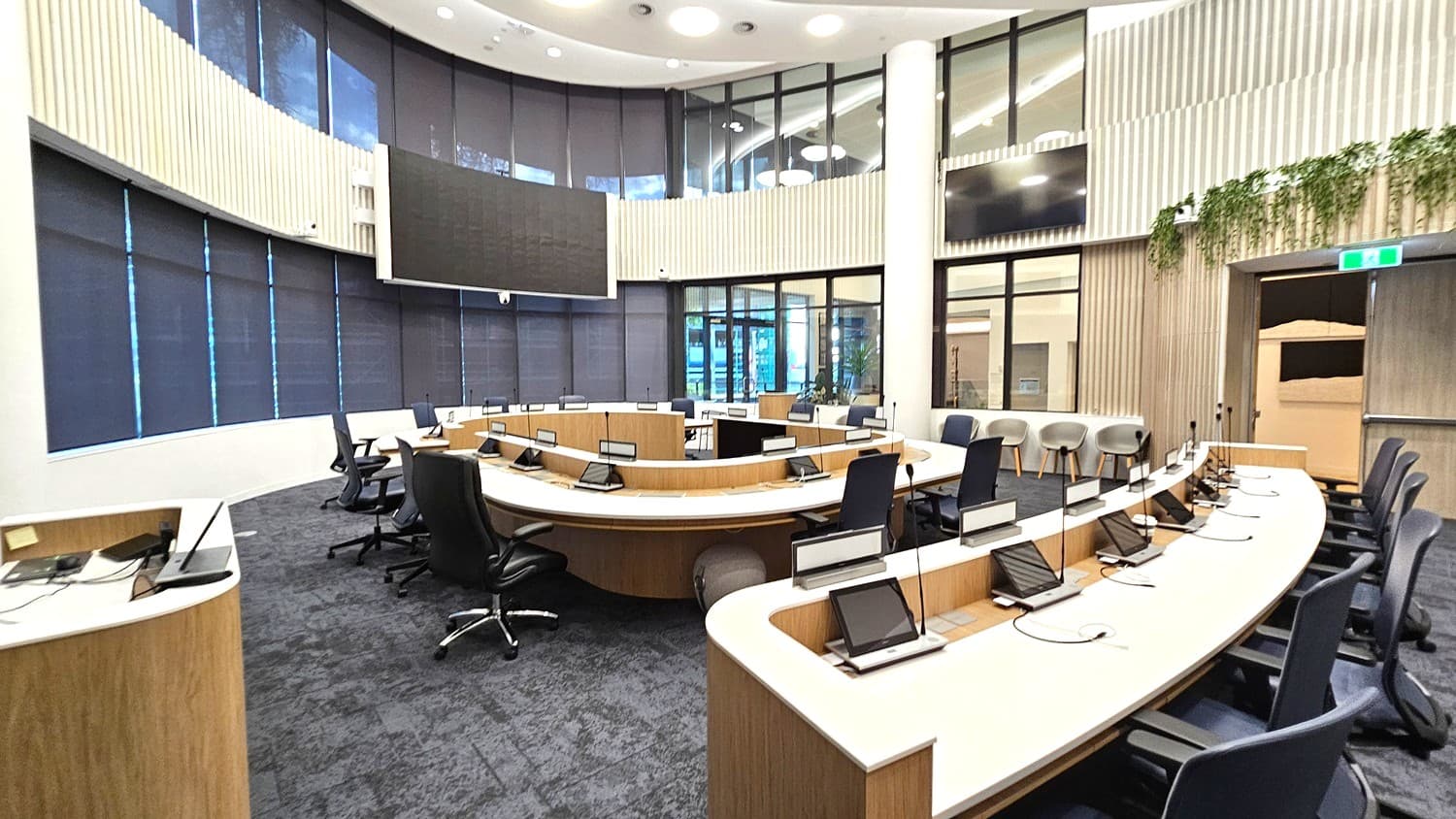 A modern conference room with a semi-circular arrangement of desks equipped with microphones and screens. Black office chairs are positioned around the desks. The floor is carpeted in a dark grey tone. Large windows with dark blinds line the curved wall in the background. The ceiling is high with circular lights. On the right, there are additional chairs and a wall with vertical wooden panelling and some greenery. A large screen is mounted on the wall.