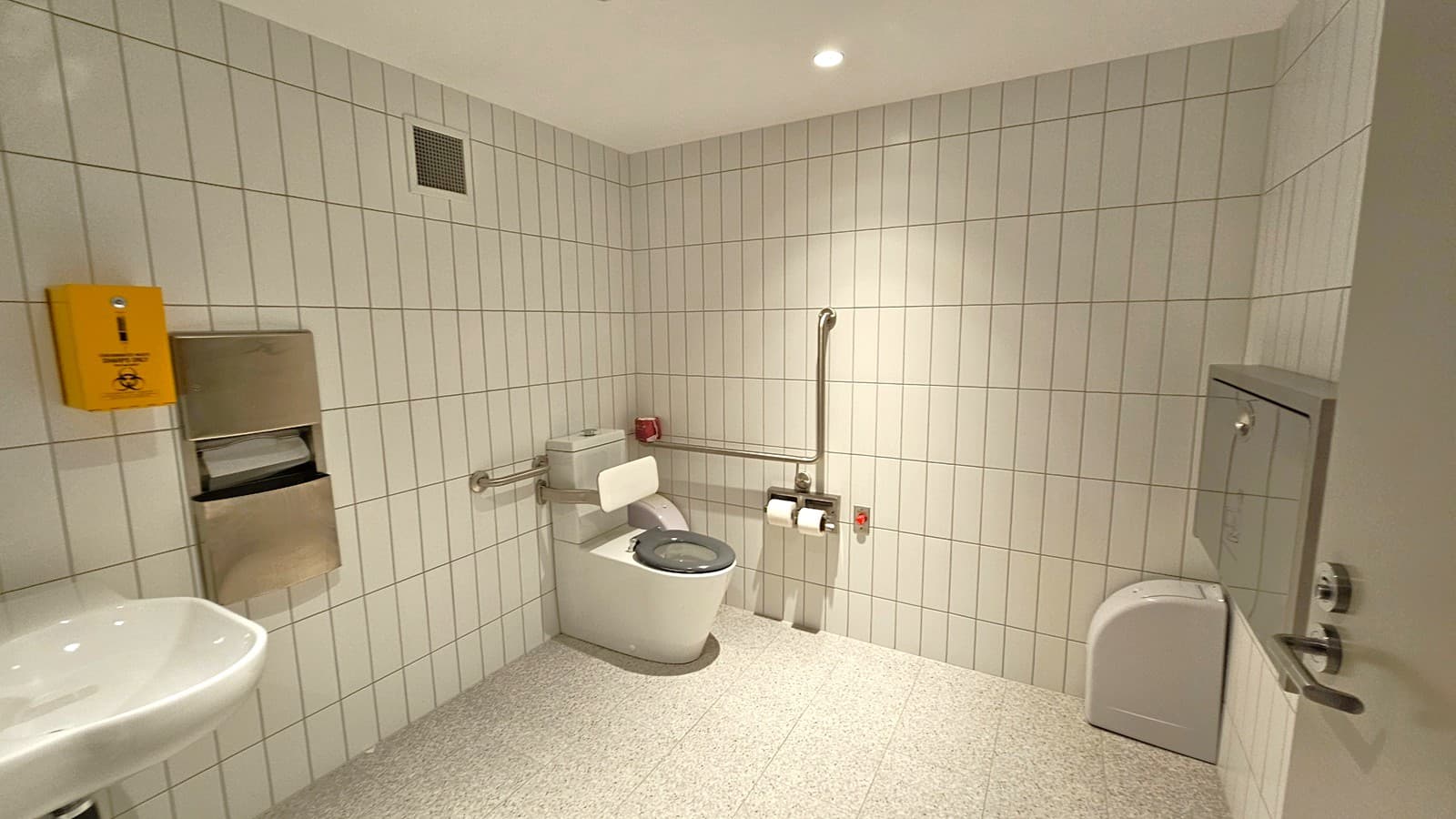 A public restroom with white tiled walls and a speckled grey floor. In the centre, a toilet with a green seat is equipped with support rails. To the right, a sanitary disposal bin is mounted on the wall. On the left, a sink is partially visible, along with a stainless steel paper towel dispenser and a yellow biohazard disposal box. The ceiling is plain with recessed lighting.