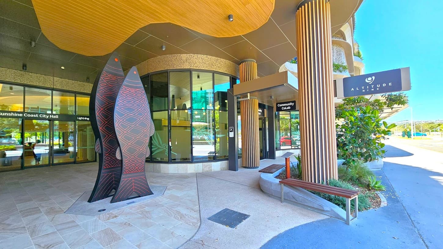 Entrance area with a modern design. On the left, there are large glass doors with signage for "Sunshine Coast City Hall" and "Lightning." In the centre, a tall, artistic sculpture resembling fish with geometric patterns stands prominently. On the right, a sign for "Altitude Nine" is visible above a wooden bench and potted plants. The ceiling features a wooden panel design, and the floor is a mix of stone and concrete. The background shows a street and distant landscape.