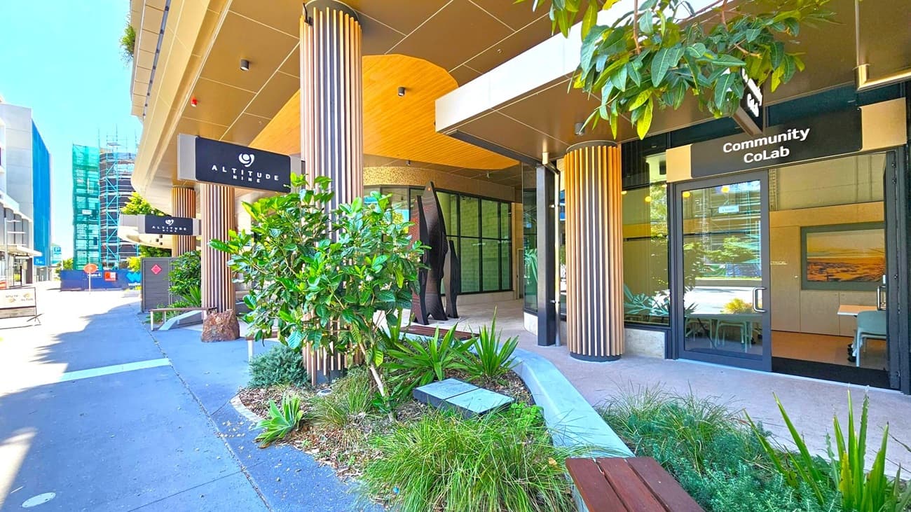 Entrance to a modern building with a sign reading "Altitude Nine" on the left and "Community CoLab" on the right. The facade features vertical wooden slats and large glass windows. A small garden with various green plants and a bench is in the foreground. The pavement is grey, and there are construction barriers and scaffolding visible in the background on the left. The ceiling is smooth and light-coloured, complementing the building's contemporary design.