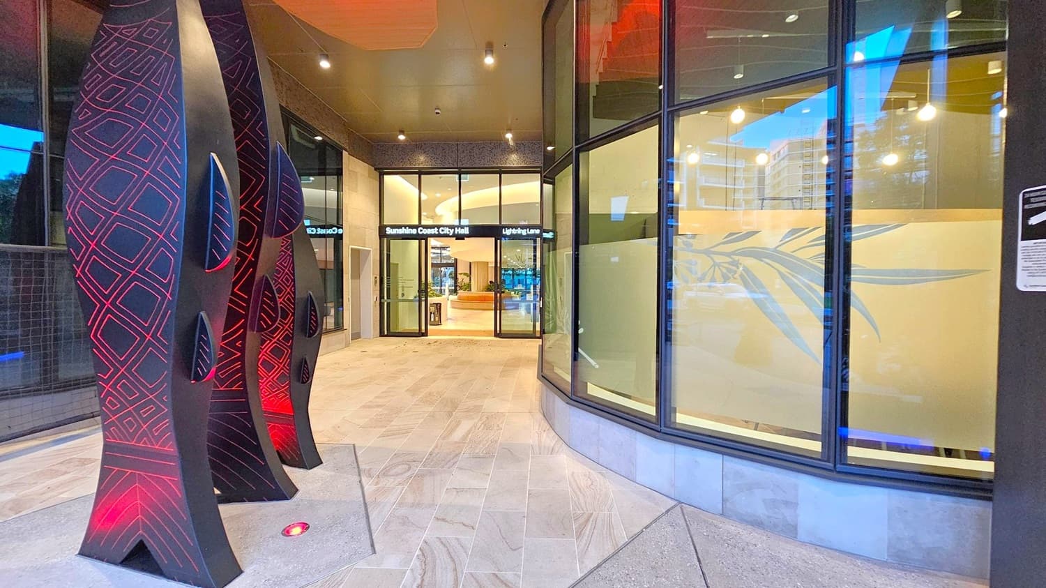 On the left, there are tall, black sculptures with red geometric patterns and small cut-out shapes. The floor is tiled with light-coloured stone. In the centre, glass doors lead to the Sunshine Coast City Hall, with a sign above reading "Lightning Lane". The interior is visible with a circular seating area and plants. On the right, large windows with frosted glass feature a leaf design. The ceiling is dark with recessed lighting.
