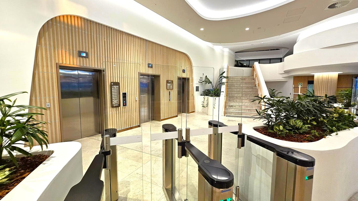 A modern interior with two lifts on the left, set in a wall with vertical wooden panelling. In the centre, a glass barrier with turnstiles. On the right, a staircase with a wooden handrail leads upwards. The foreground features planters with green plants, and the ceiling is smooth with recessed lighting. The floor is light-coloured and polished.