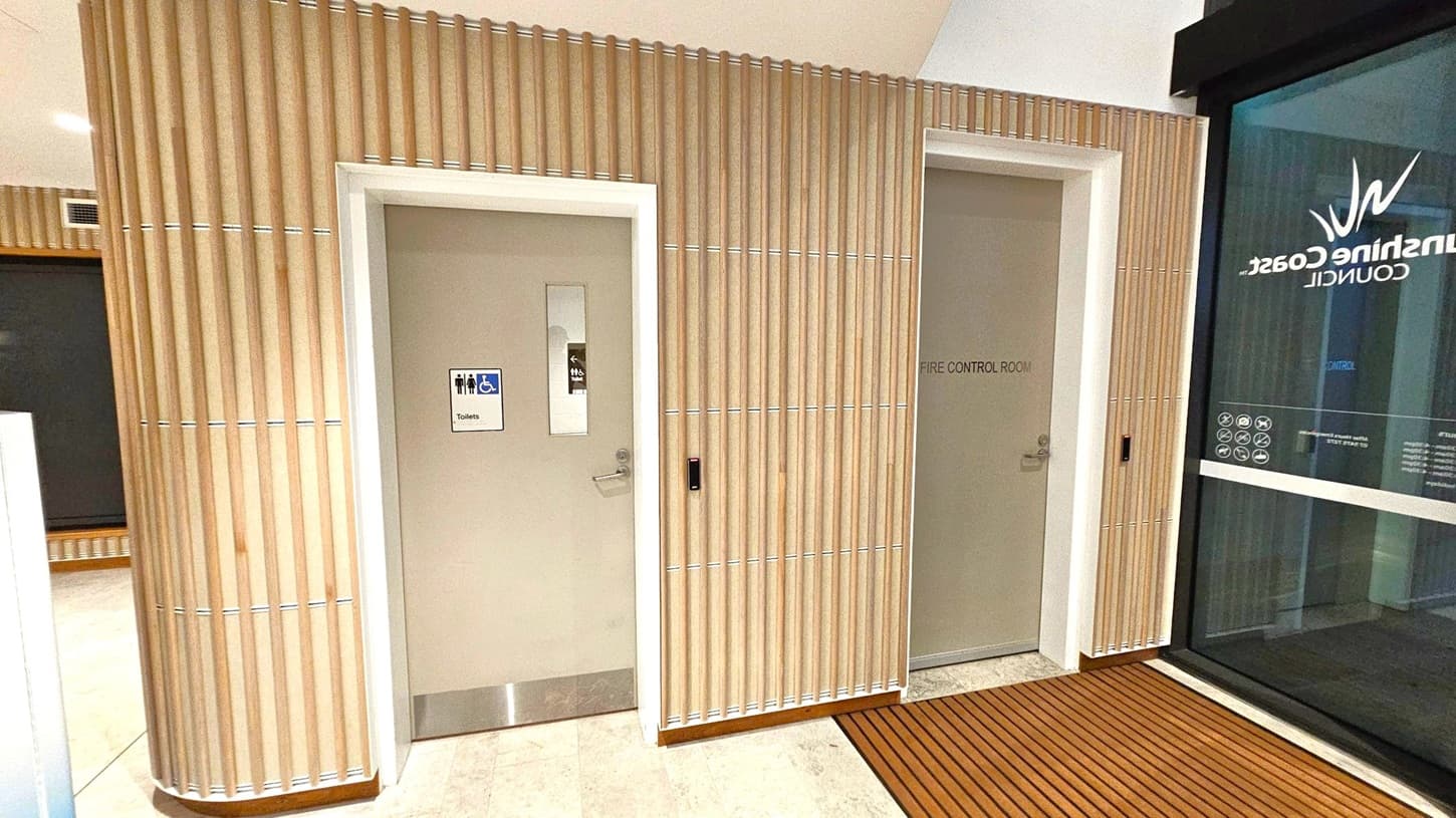 A hallway with two doors on a wall featuring vertical wooden slats. The left door has a sign indicating toilets, including accessible facilities, and a small window. The right door is labelled "Fire Control Room." To the right, a glass panel displays the words "Sunshine Coast Council" and various symbols. The floor is tiled, transitioning to a wooden slat mat near the glass panel. The ceiling is plain with recessed lighting.