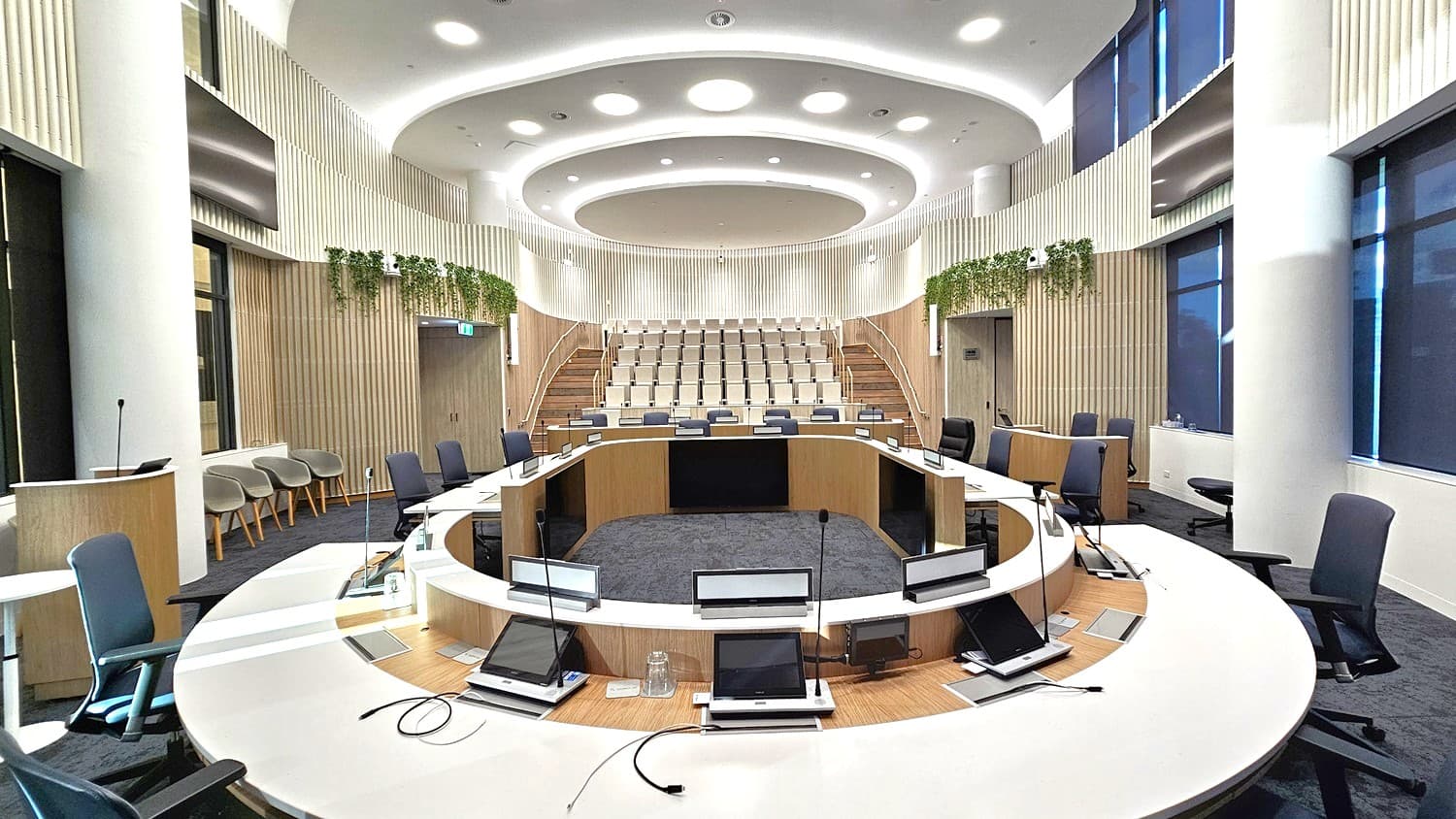 Wide angle chamber view.  Council Chambers.  Sunshine Coast City Hall.  A modern conference room with a circular seating arrangement. In the foreground, there are several chairs and desks equipped with microphones and screens. The middle features a central circular area with additional seating. The background shows tiered seating with light-coloured chairs. The walls are adorned with vertical wooden panels and greenery. The ceiling has circular light fixtures.
