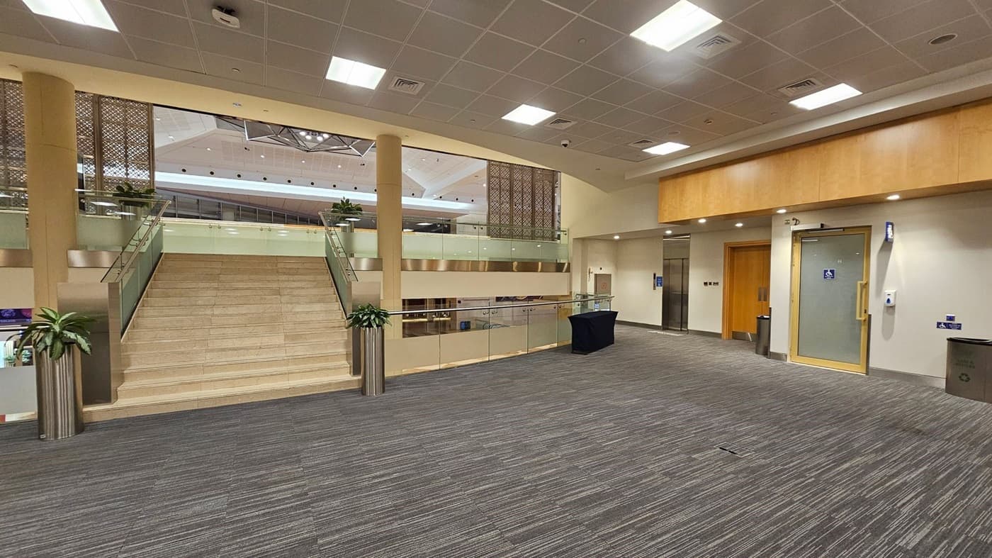 A spacious indoor area with a wide staircase in the centre, flanked by glass railings and potted plants. The floor is covered with grey and green striped carpet. On the right, there is a wooden door and a glass door with a disabled access sign. The ceiling is fitted with square lights and a grid pattern. In the background, there are decorative panels and a glimpse of an upper level with more plants.
