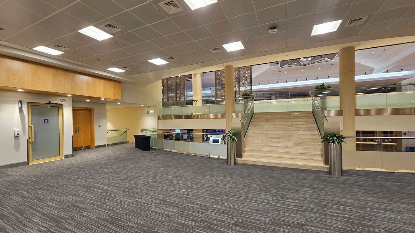 A spacious indoor area with a grey carpeted floor. On the left, there is a door with a disabled access sign and a wooden double door. In the centre, a wide staircase with glass railings leads to an upper level. Potted plants are placed at the base and top of the stairs. The ceiling is fitted with square lights and ventilation panels. The upper level has a decorative panel and overlooks a lower area with visible signage.