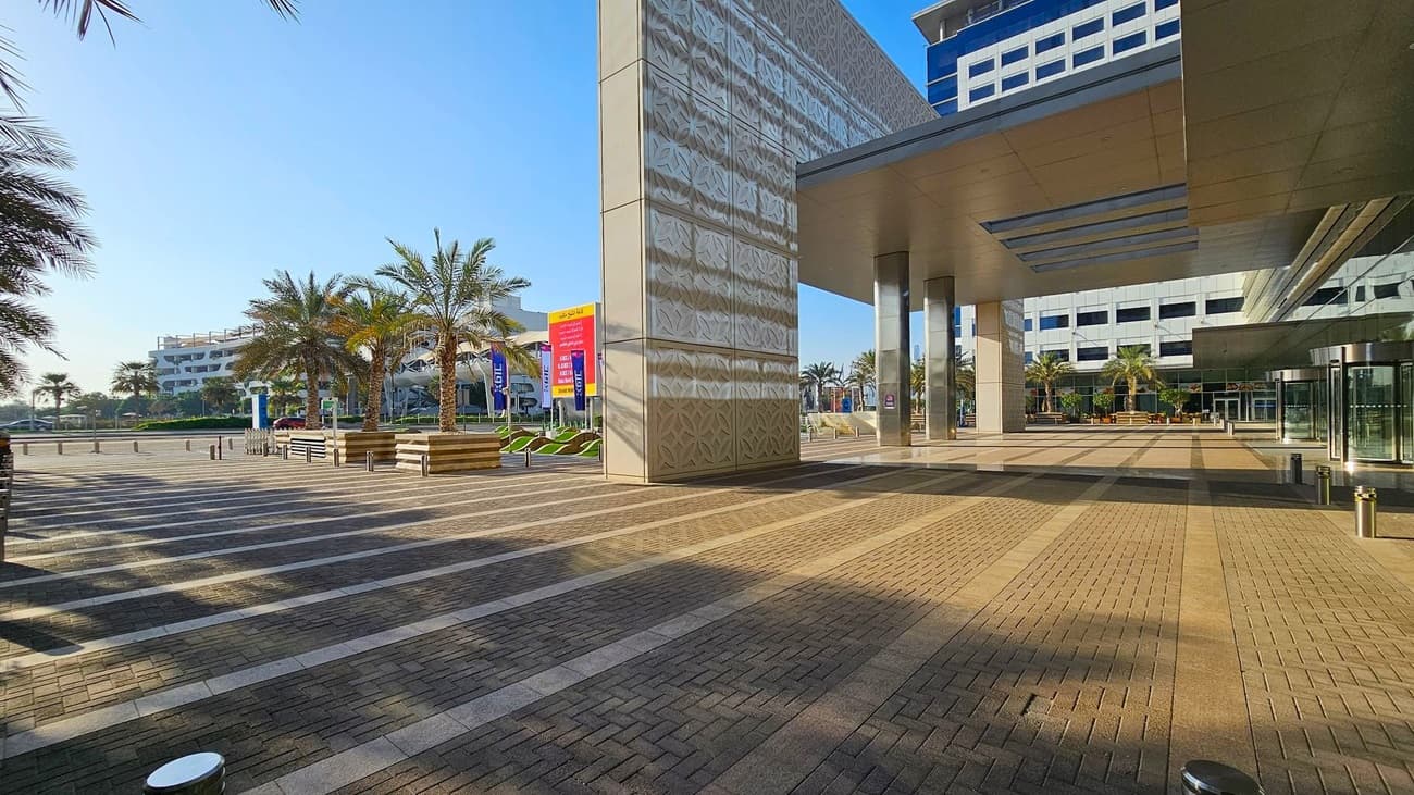 A modern outdoor plaza with a patterned, reflective building on the right. The ground is paved with alternating light and dark bricks. Palm trees and benches are on the left, with a large, colourful sign in the background. The area is open and spacious, with a clear blue sky above. A glass entrance with revolving doors is visible on the right side of the building.
