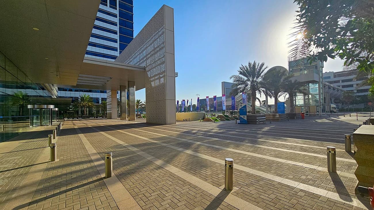 A modern urban plaza with a patterned pavement and metal bollards. On the left, a tall building with reflective glass windows and a covered walkway. In the centre, palm trees and a series of flags line the path. On the right, a structure with glass and metal elements, partially obscured by trees. The sun is visible through the trees, casting shadows on the ground.
