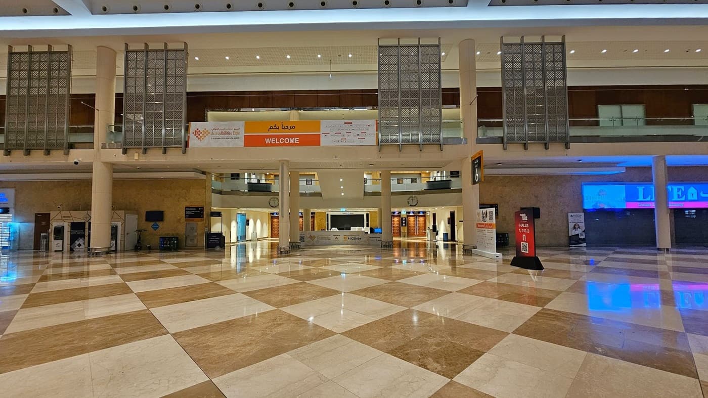 A spacious indoor hall with a polished, reflective floor featuring a diamond pattern in shades of beige and brown. On the left, there are signs for Hall 6 and Forex Expo, along with a clock and elevators. The right side has a "Super Sale" sign and various illuminated displays. The ceiling is high with geometric designs and bright lighting. The area is lined with shops and advertisements, and there are few people in the distance.