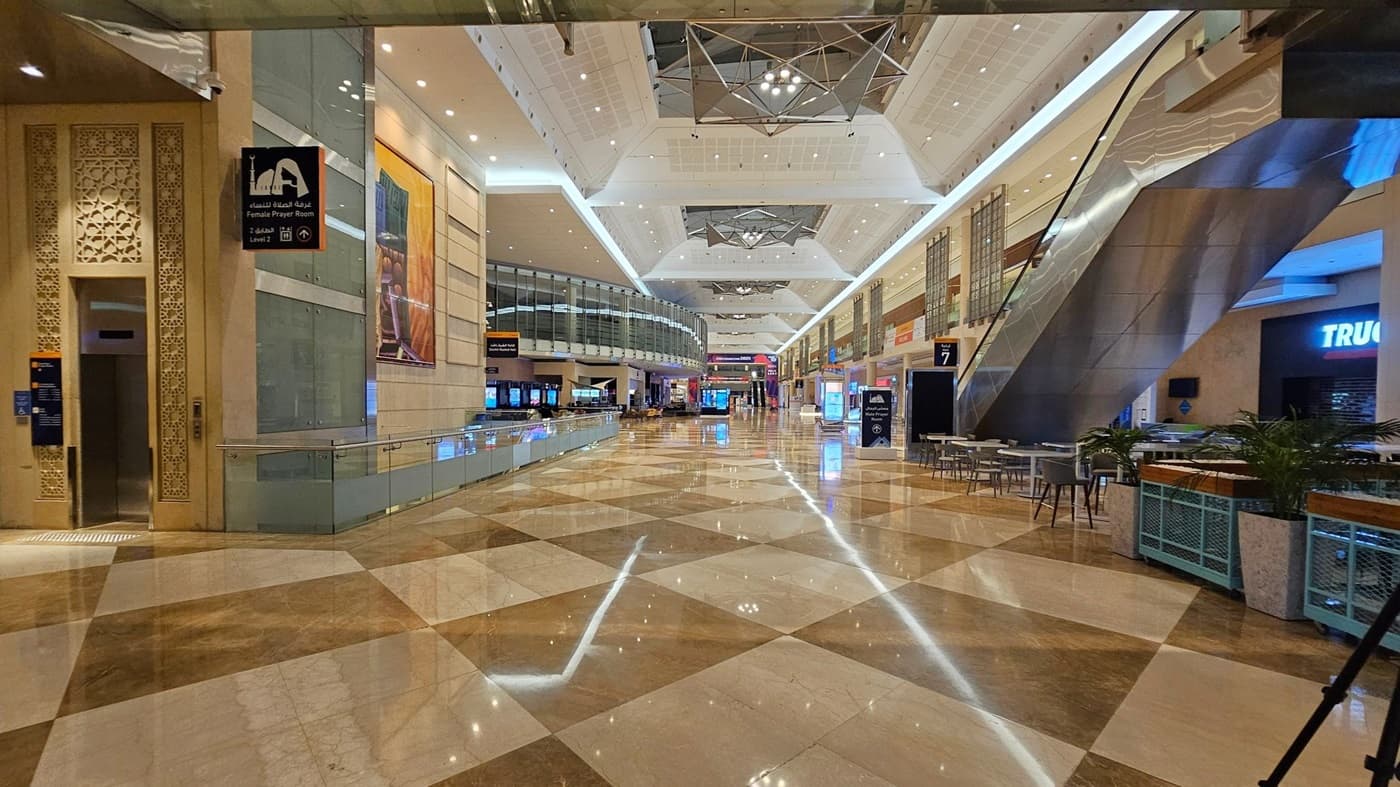 A spacious indoor area with a polished, reflective floor featuring a geometric pattern. On the left, there's a sign for a female prayer room and an intricately designed wall. The centre has a glass railing and a curved glass structure. On the right, there are escalators, seating areas with tables and chairs, and a shop with a partially visible sign. The ceiling is high with geometric light fixtures and recessed lighting.
