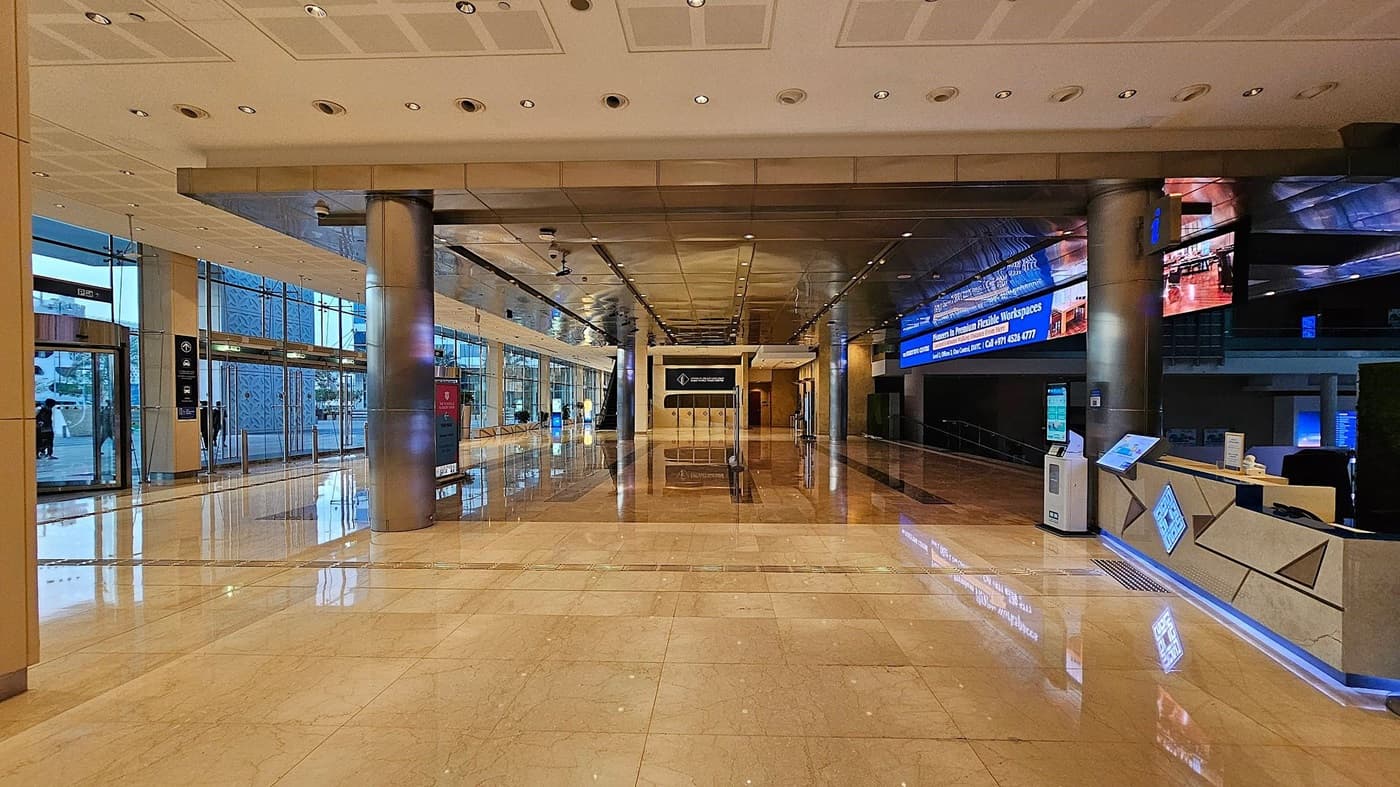 A spacious, modern lobby with a shiny marble floor and a grid-patterned ceiling with recessed lighting. On the left, there are dark reflective surfaces and a reception area. The right side features large glass windows with views of an outdoor area, including palm trees and banners. In the centre, there are glass doors and a large pillar. The space is well-lit, with reflections visible on the floor