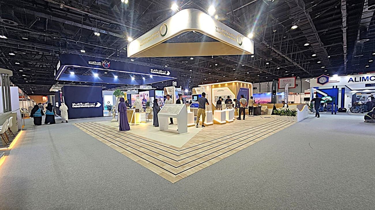A large exhibition hall with a central booth featuring a wooden floor and illuminated displays. Above, a suspended sign reads "Dubai Municipality." On the left, a group of people walk past a dark display screen. On the right, another booth with the name "ALIMCO" is visible. The ceiling is high with exposed beams and lighting fixtures. The floor is a mix of carpet and wood.