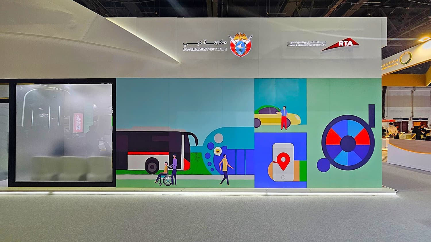 A large wall display with a colourful mural featuring transportation themes. On the left, there's a frosted glass panel with a faint reflection of an interior space. The mural includes illustrations of a bus, a person in a wheelchair, a car, and a smartphone with a location pin. Above, the logos of the Government of Dubai and the Roads & Transport Authority (RTA) are visible. The floor is grey, and the ceiling is dark with lighting fixtures.