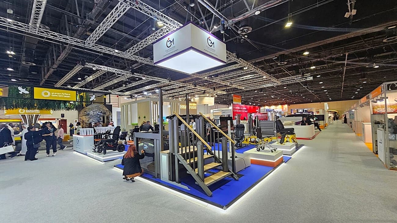 A trade show floor with various exhibits. In the centre, there's a display featuring a small staircase and platform on a blue carpet. Above, a large hanging sign with a logo. To the left, people are gathered near a booth with a green and yellow sign. On the right, more exhibits and a long aisle. The ceiling is high with exposed beams and lighting fixtures. The floor is carpeted in grey.
