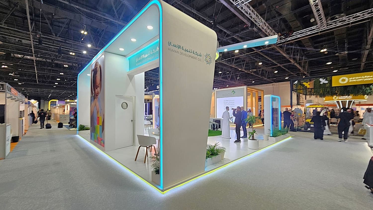 A trade show booth with a modern design featuring a white structure with blue neon lighting accents. On the left, there's a large image of a child and a small seating area with a white chair. The booth is branded with "Human Development Co." in both English and Arabic. In the background, people are walking around, and other booths are visible, including one with a yellow sign. The ceiling is high with exposed beams and lighting fixtures. The floor is carpeted in grey.