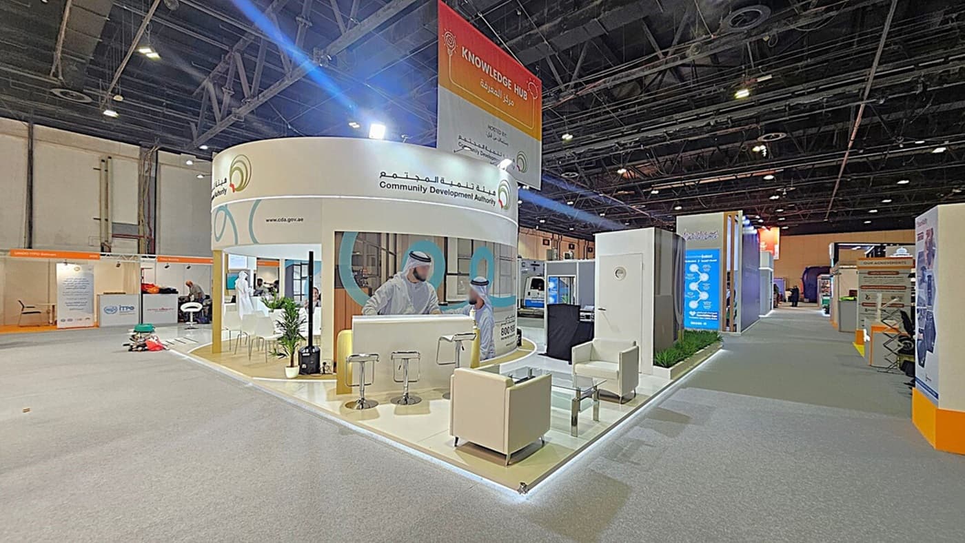 A trade show booth with a circular design, featuring the Community Development Authority. The booth includes a counter with stools, a glass table, and white armchairs. A large poster with two men in traditional attire is displayed. The floor is carpeted in grey, and the ceiling has exposed beams and lighting. Adjacent booths and informational displays are visible along the aisle. A "Knowledge Hub" sign hangs above.
